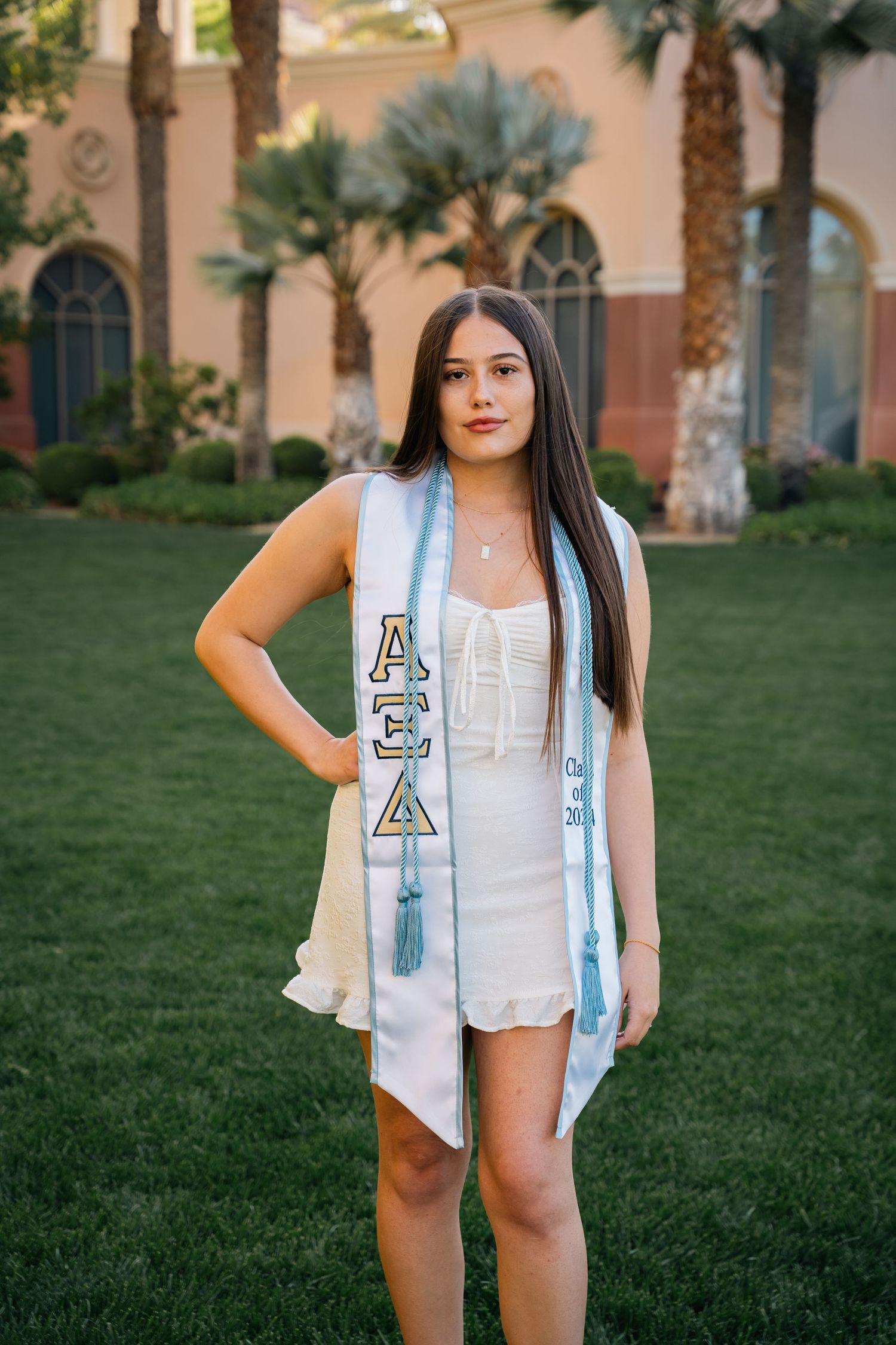Graduate wearing white dress and decorated sorority stole stands on lawn with palm trees in background.
