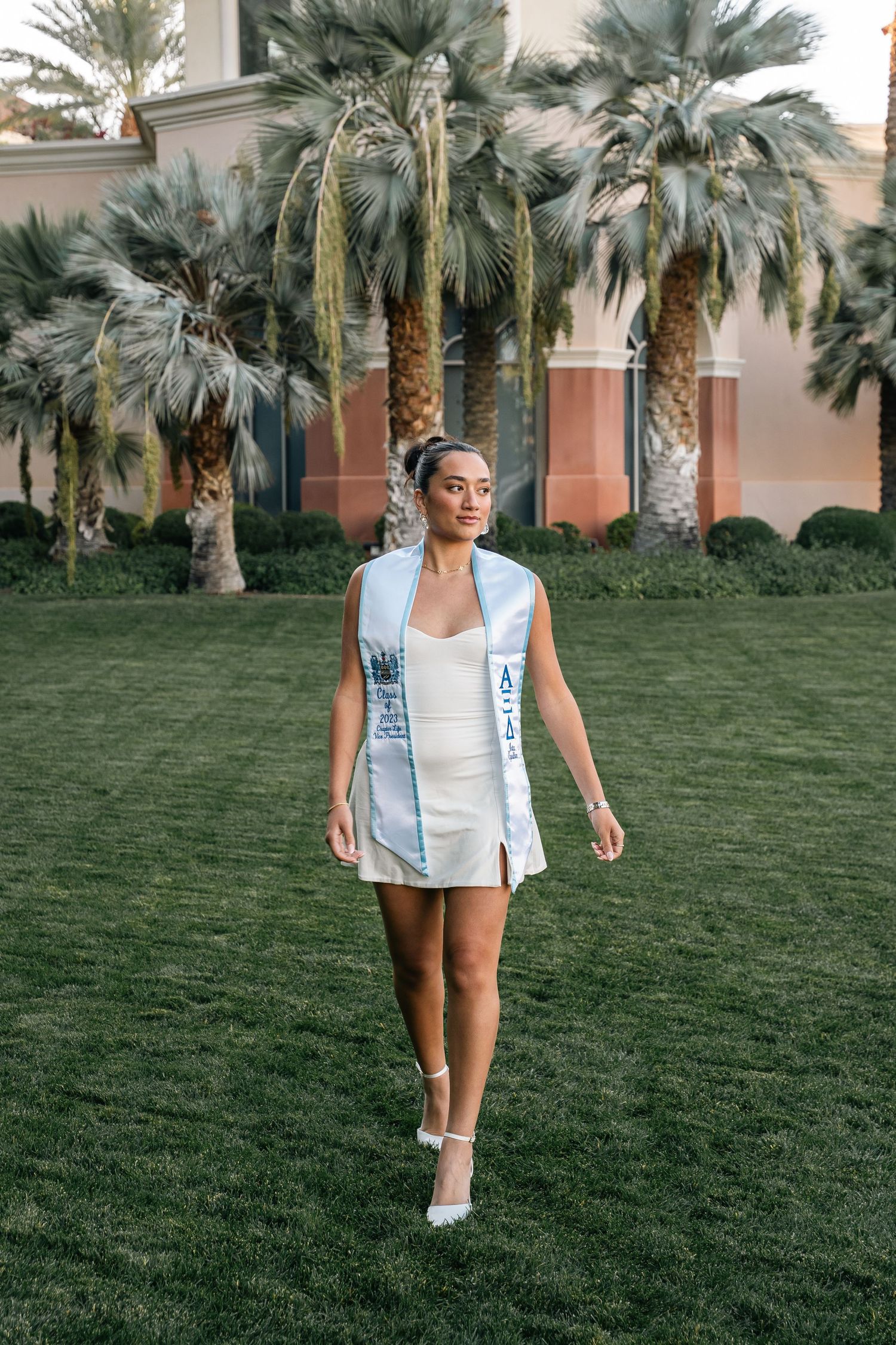 Person in white dress and blue sash walking on green lawn with palm trees at sunset.
