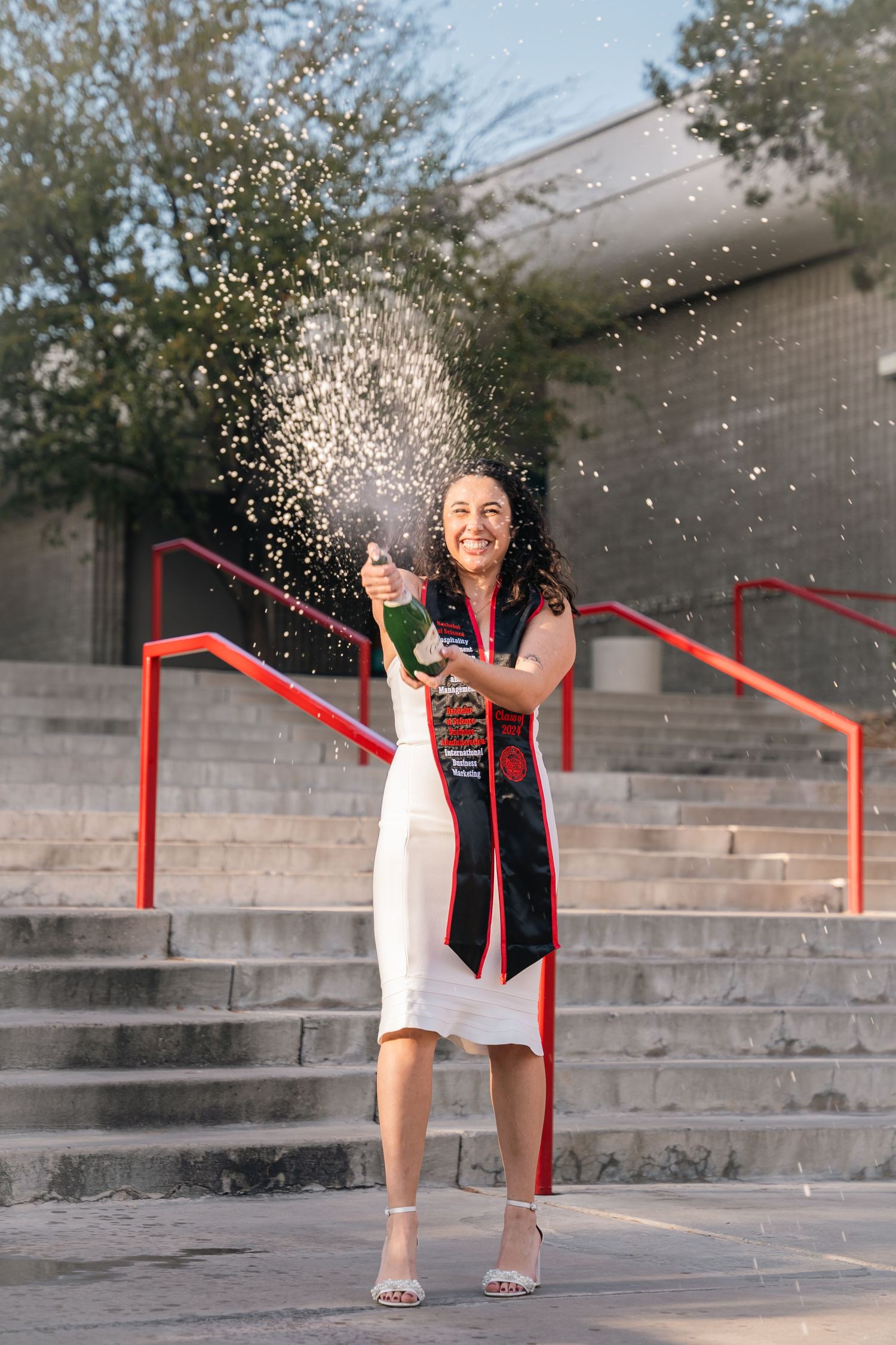 Graduate in academic regalia pops champagne on concrete steps creating celebratory spray.