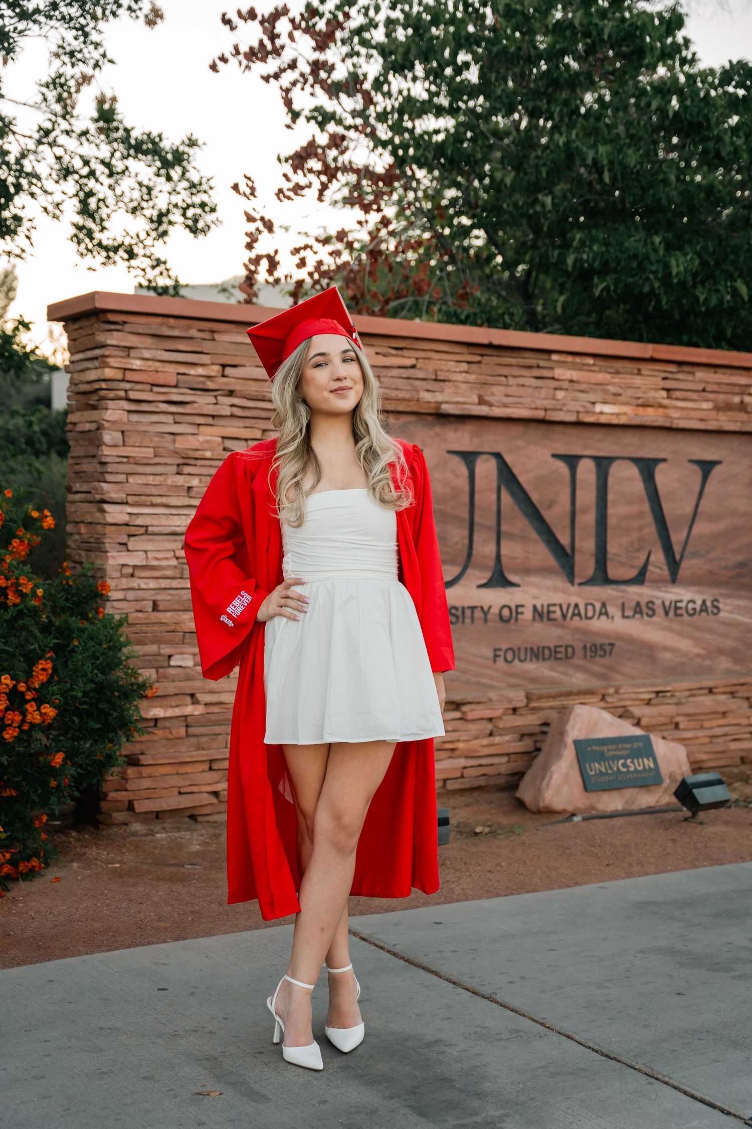 Graduate poses in red cap and gown against UNLV stone wall sign during graduation photoshoot.