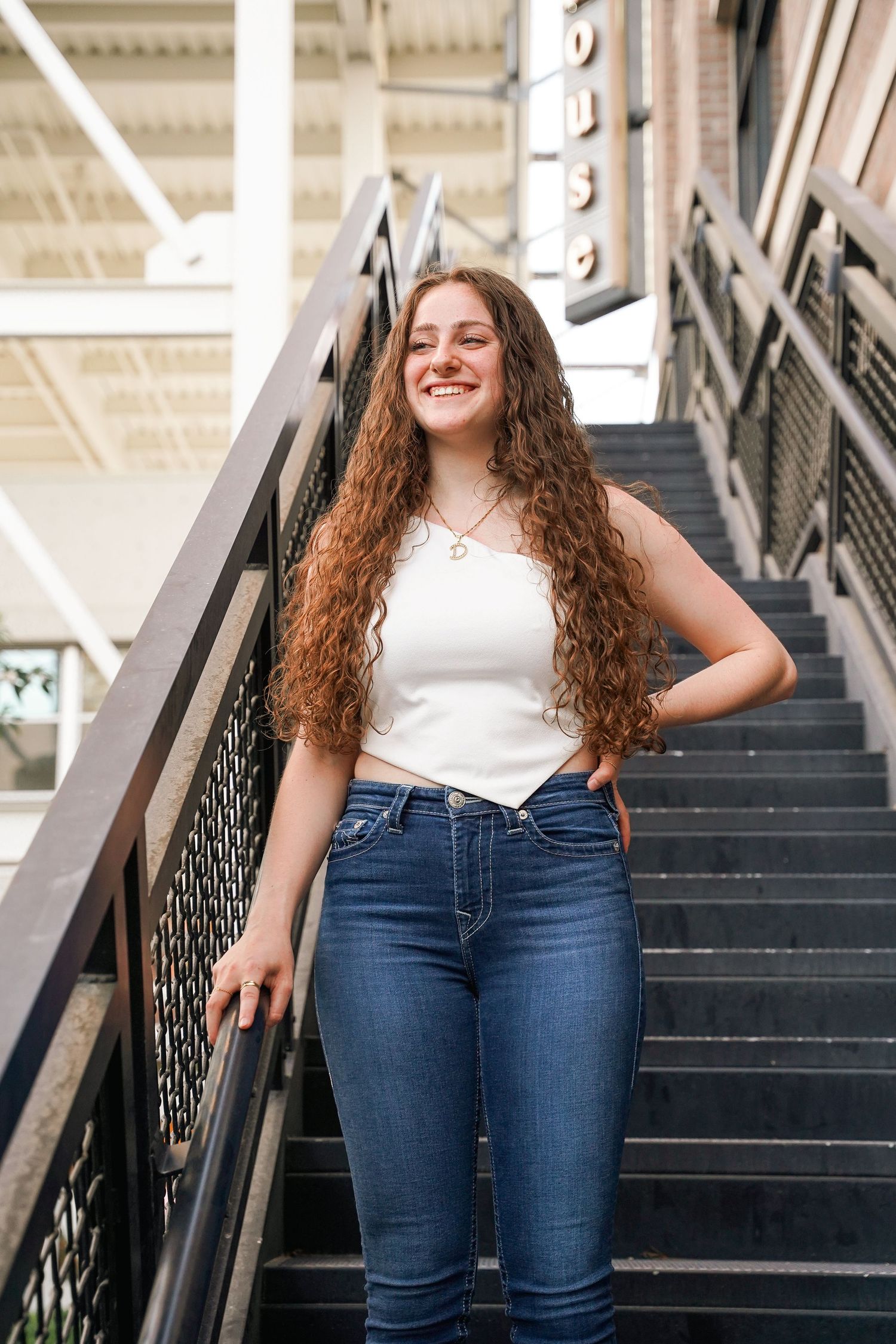 Graduate with long wavy hair poses on industrial staircase in casual attire and graduation cap.