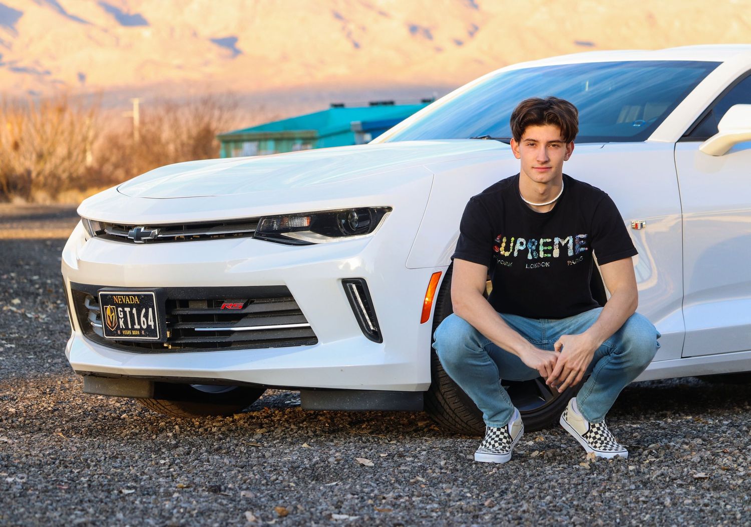 Person in black t-shirt sits casually in front of white Chevrolet Camaro at sunset.