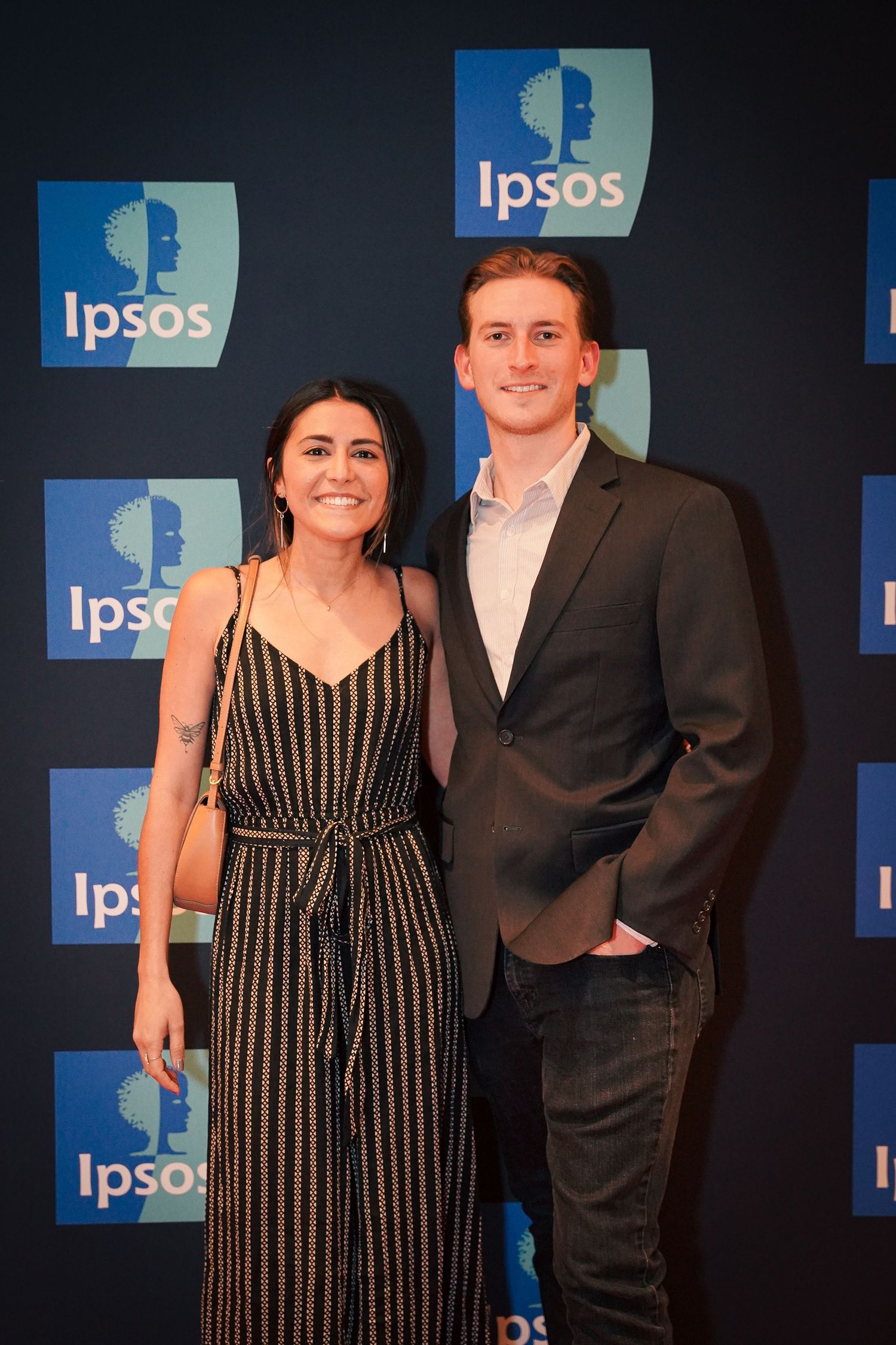 A pair pose in formal attire at an Ipsos event with blue branding and dark background.