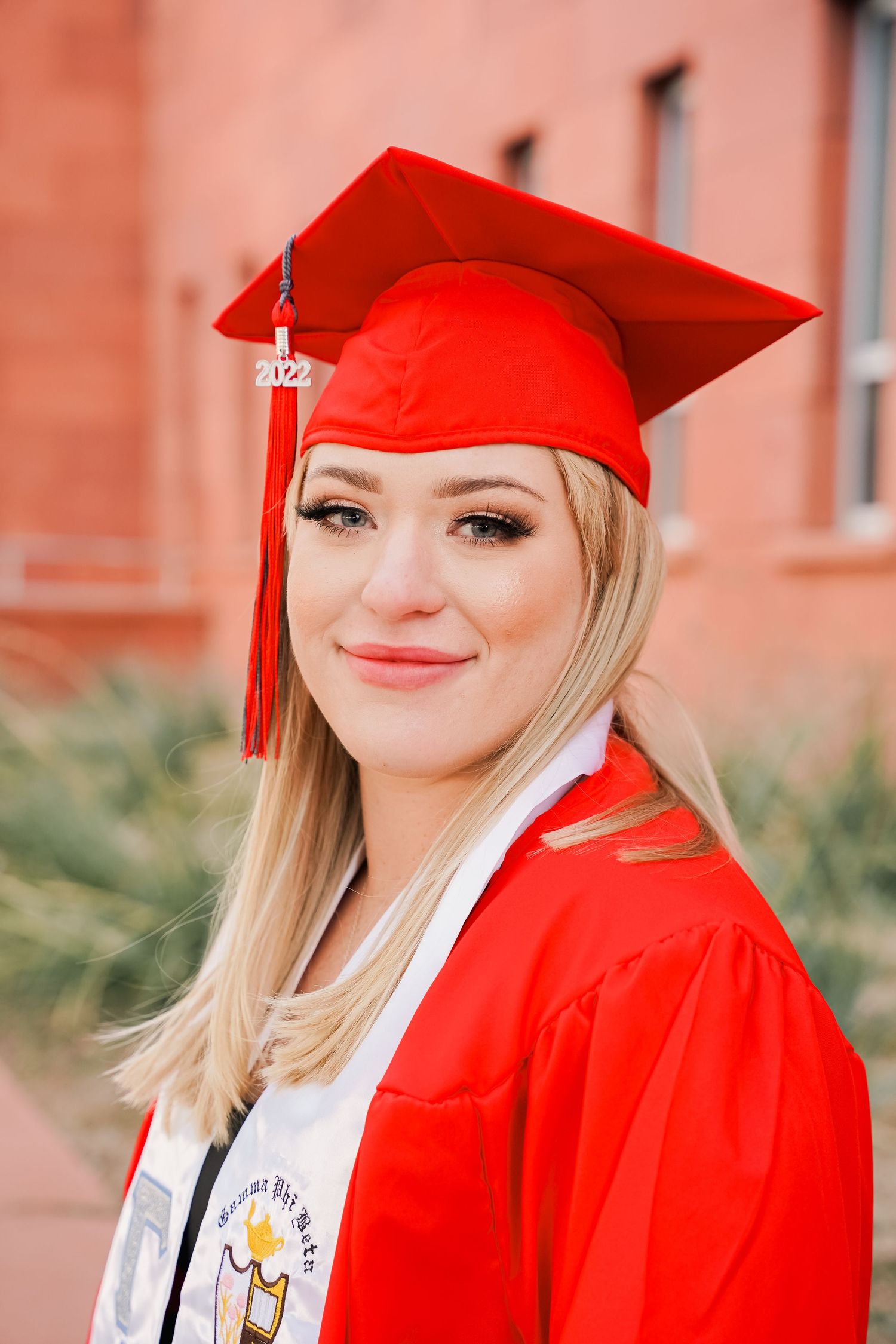 Graduate wearing bright red graduation regalia and honor stole poses against orange wall.