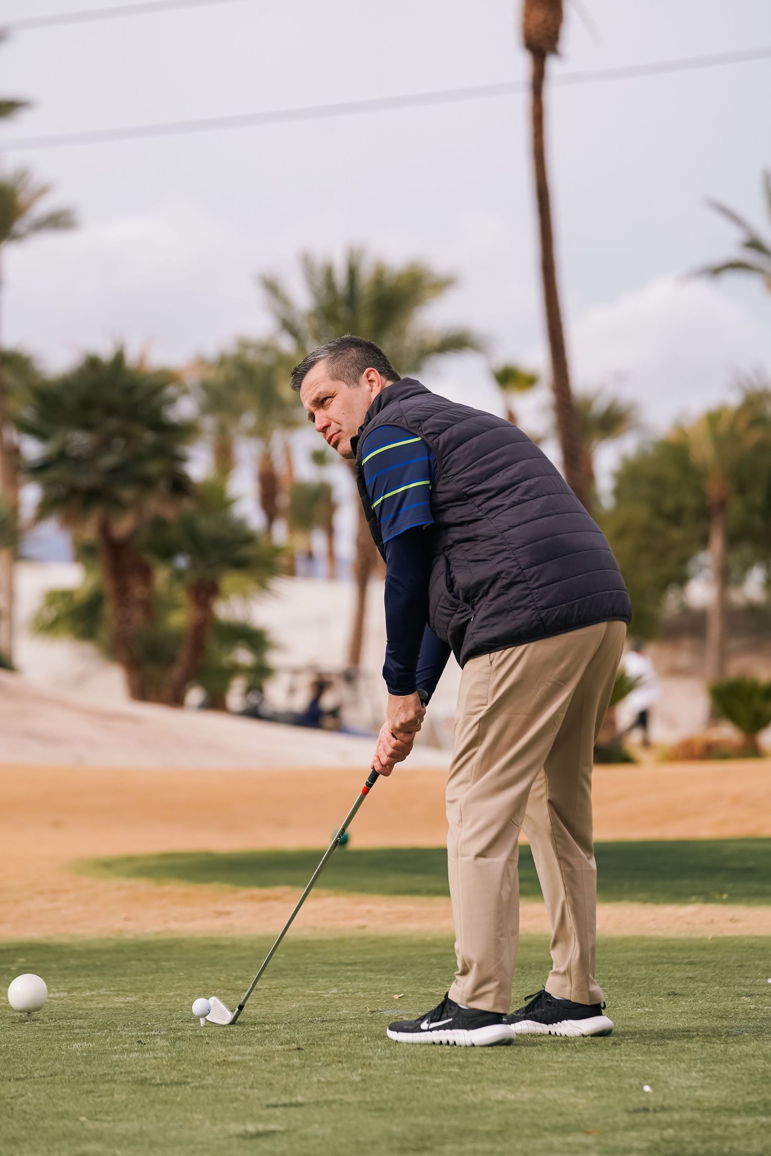 Golfer in dark jacket and khakis takes swing on desert golf course surrounded by palm trees.