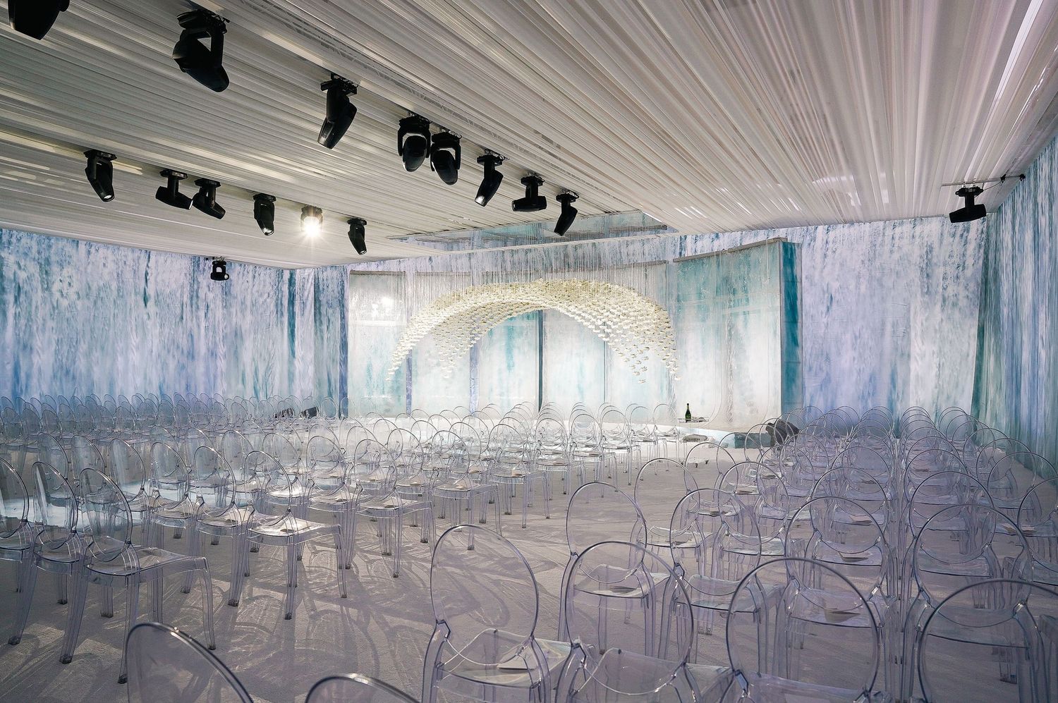 Modern wedding ceremony setup with clear chairs and illuminated floral arch in ice-blue themed space.