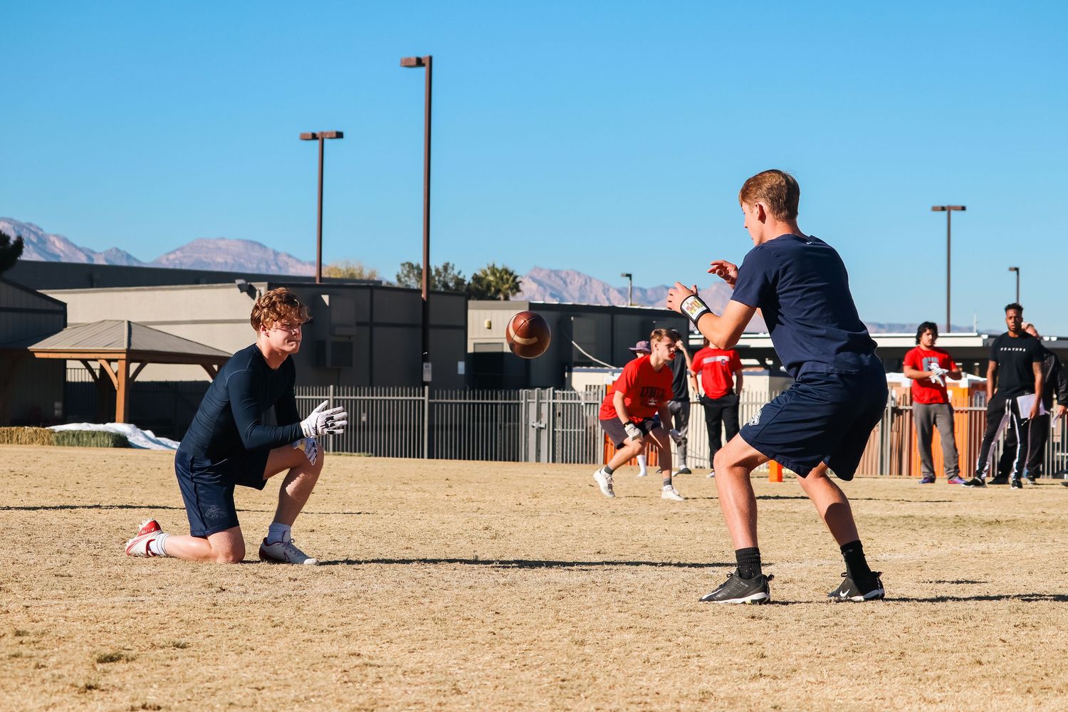 Athletes practice football drills on a dirt field during daytime.