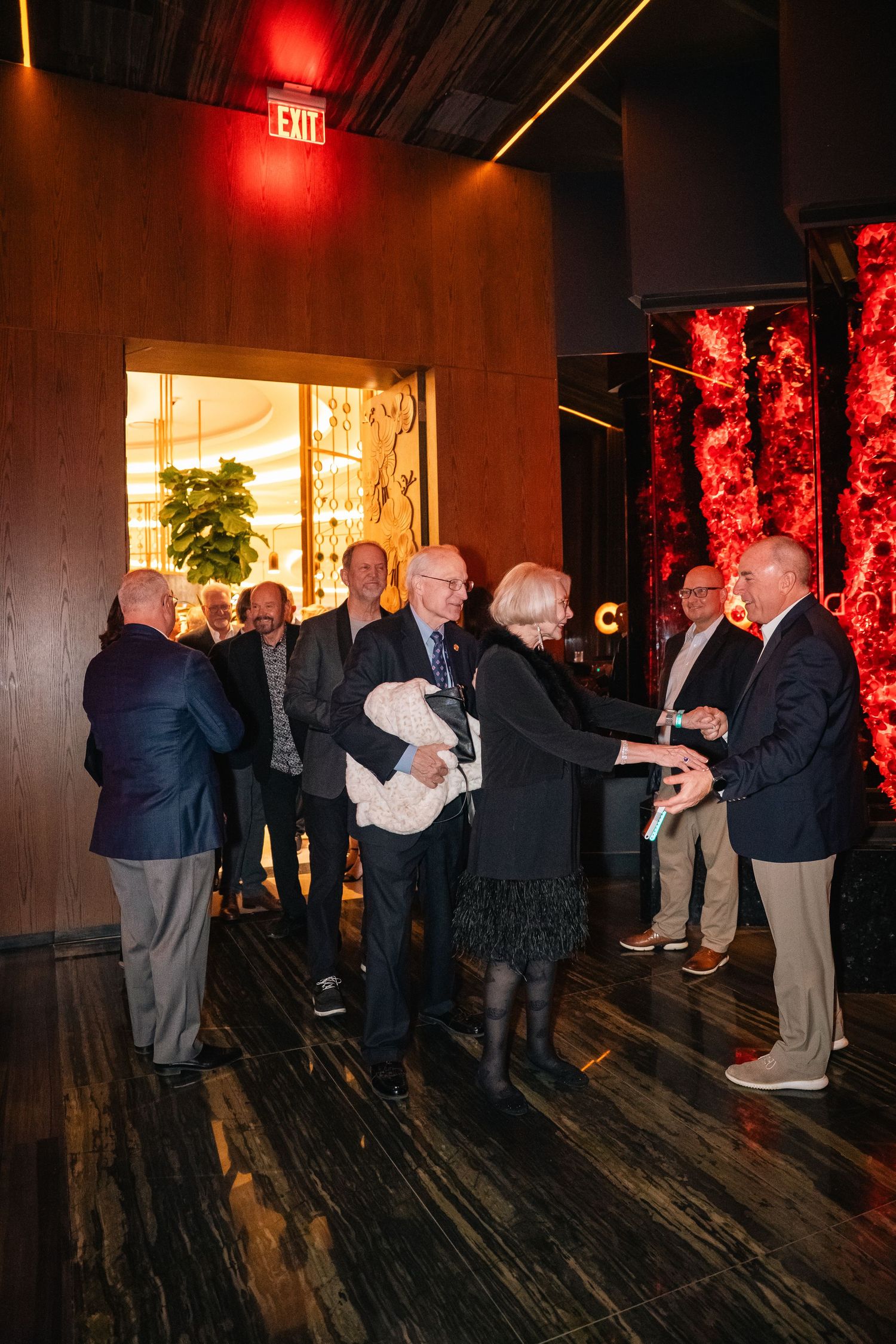 A group of guests gather in a dimly lit venue with wood floors and red lighting for a social event.