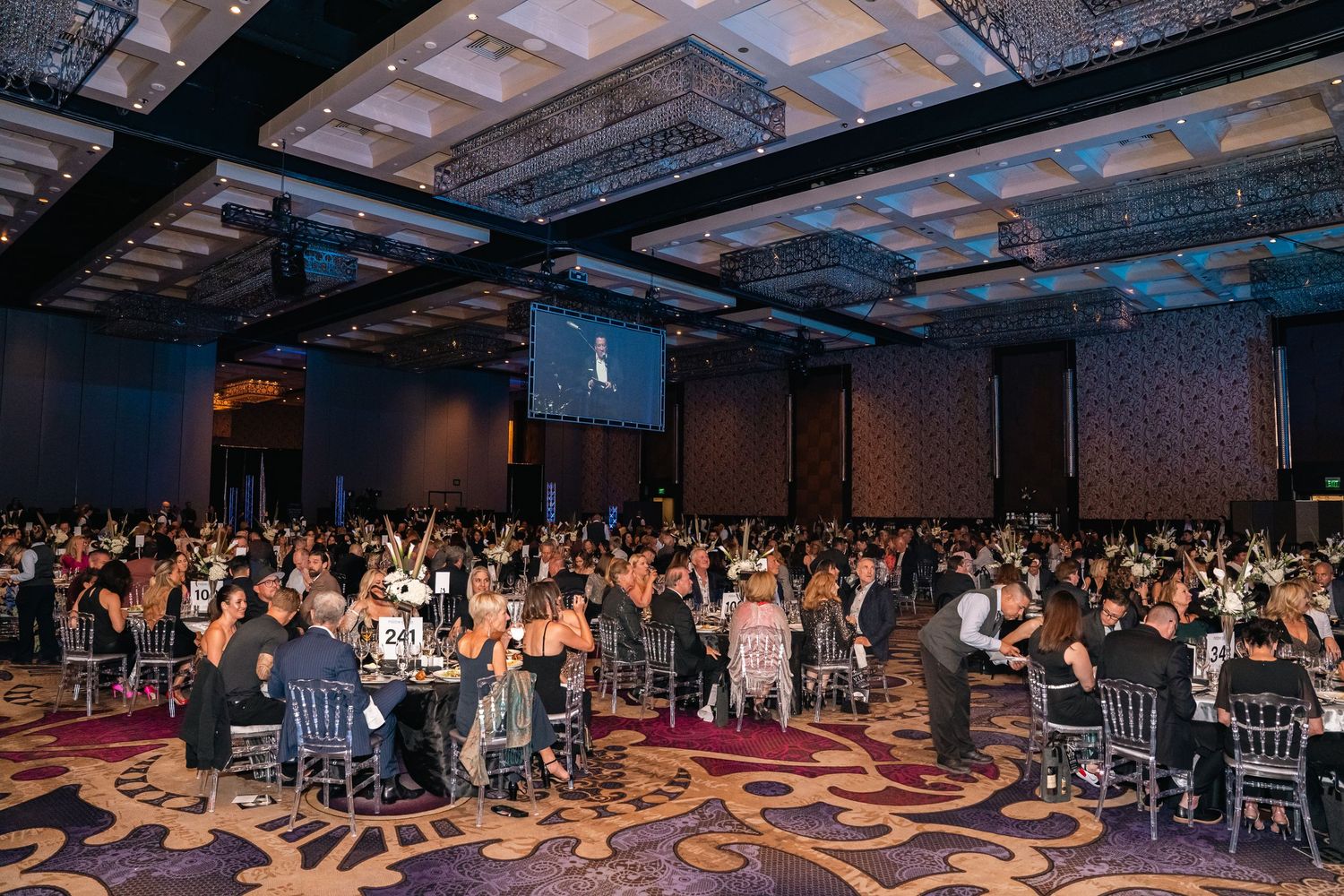 Large formal banquet hall filled with guests at round tables during an evening event.