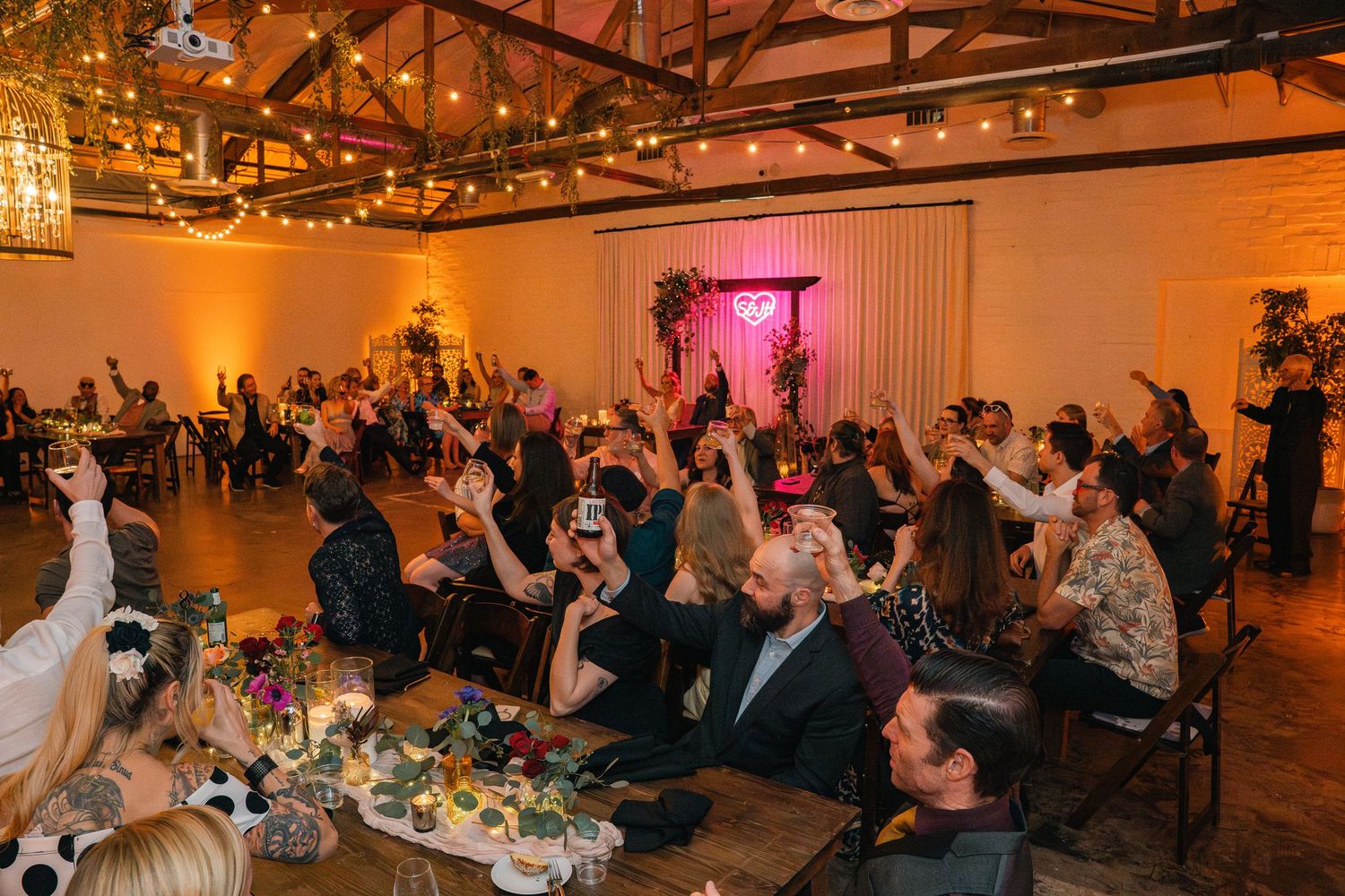Wedding reception guests raise their hands in celebration under warm lighting and exposed beams.