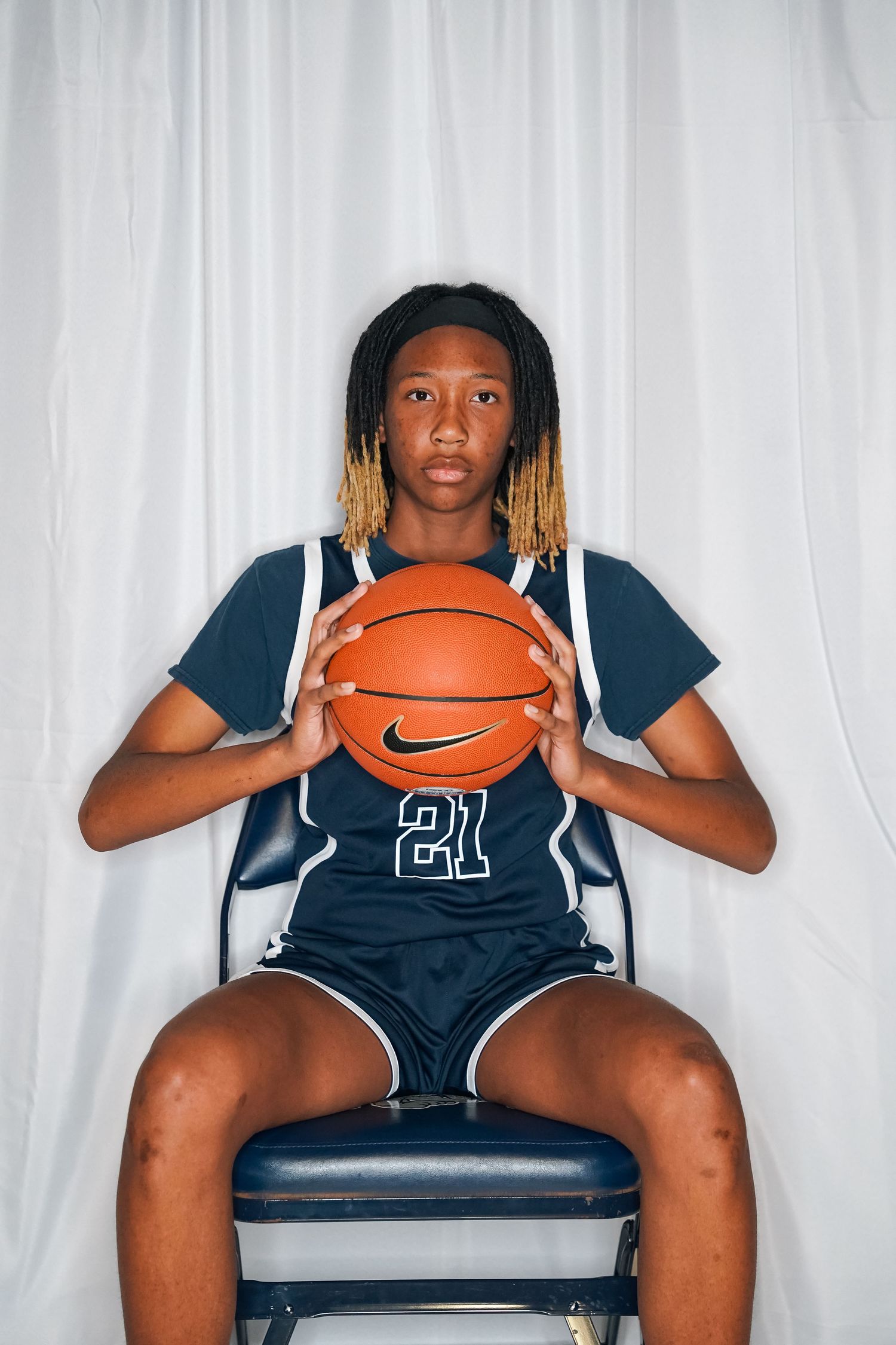 Basketball player in navy jersey number 21 sits holding ball during team photoshoot.
