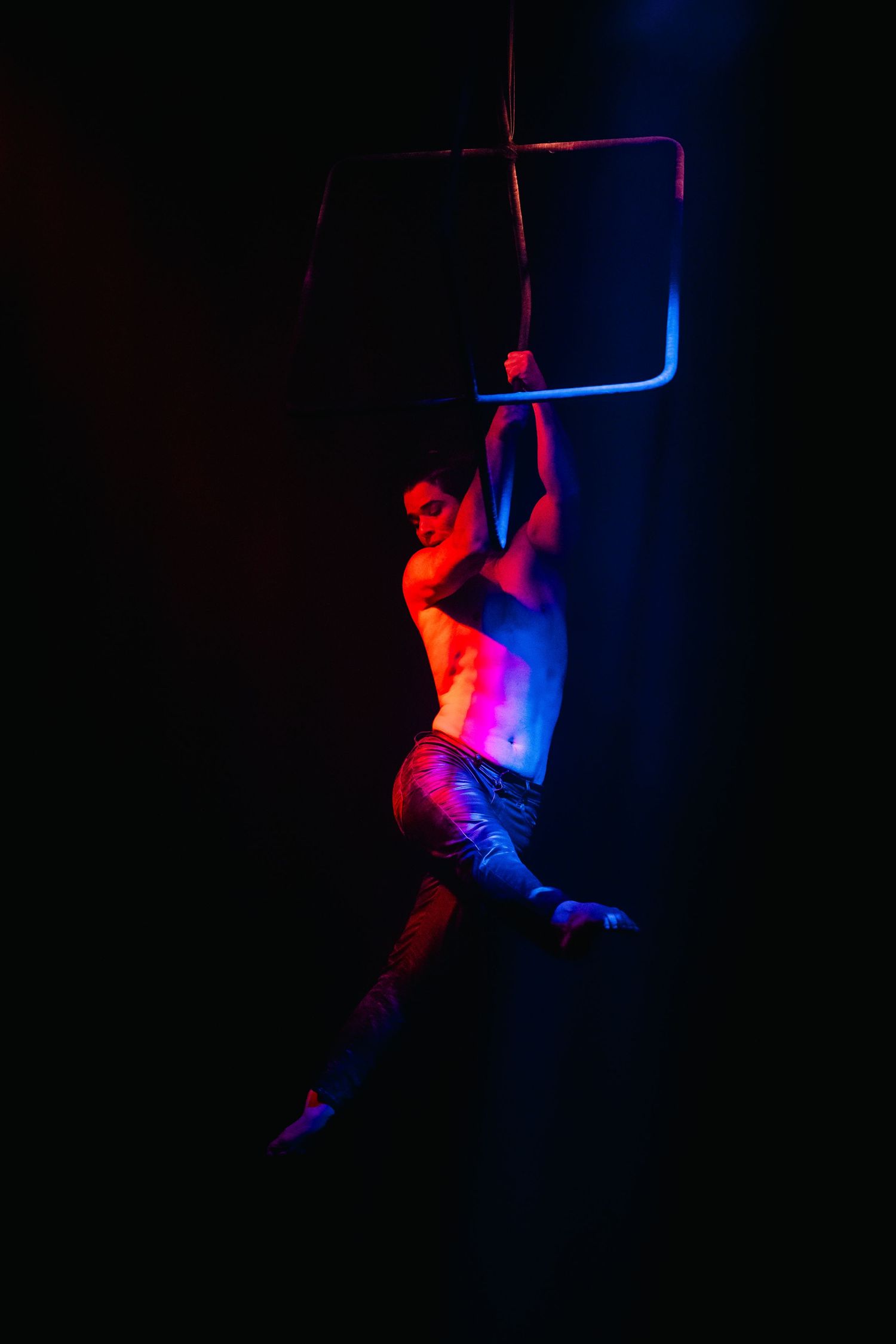 Aerial performer hanging from geometric prop in dramatic red and blue lighting.