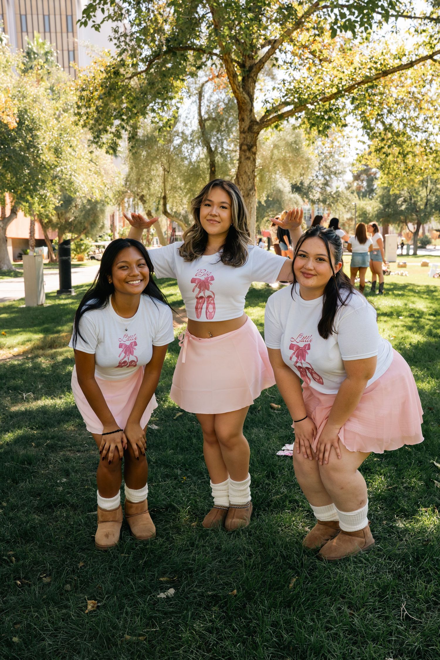 Three friends in matching pink skirts and white t-shirts with knee socks pose on grass.