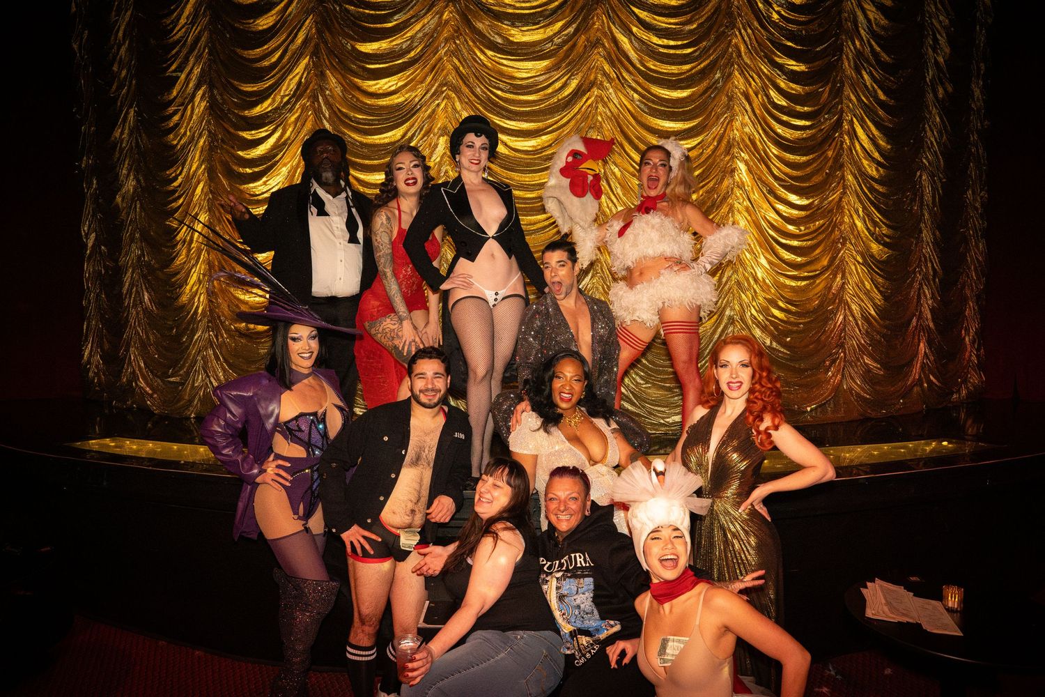 Theatrical performers posing together on stage in front of gold curtain.
