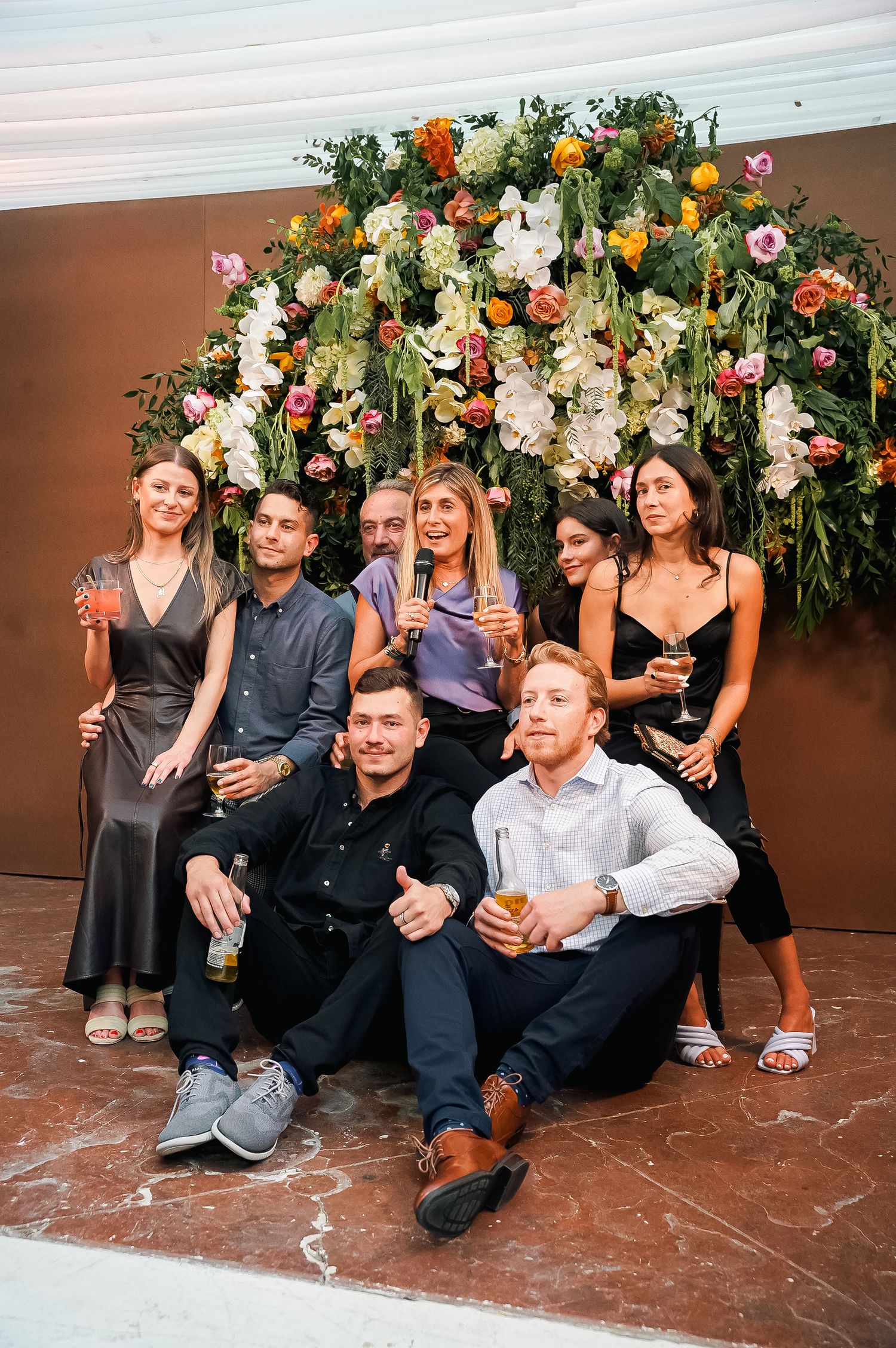 Group poses in front of a floral arch with colorful blooms during an elegant outdoor celebration.