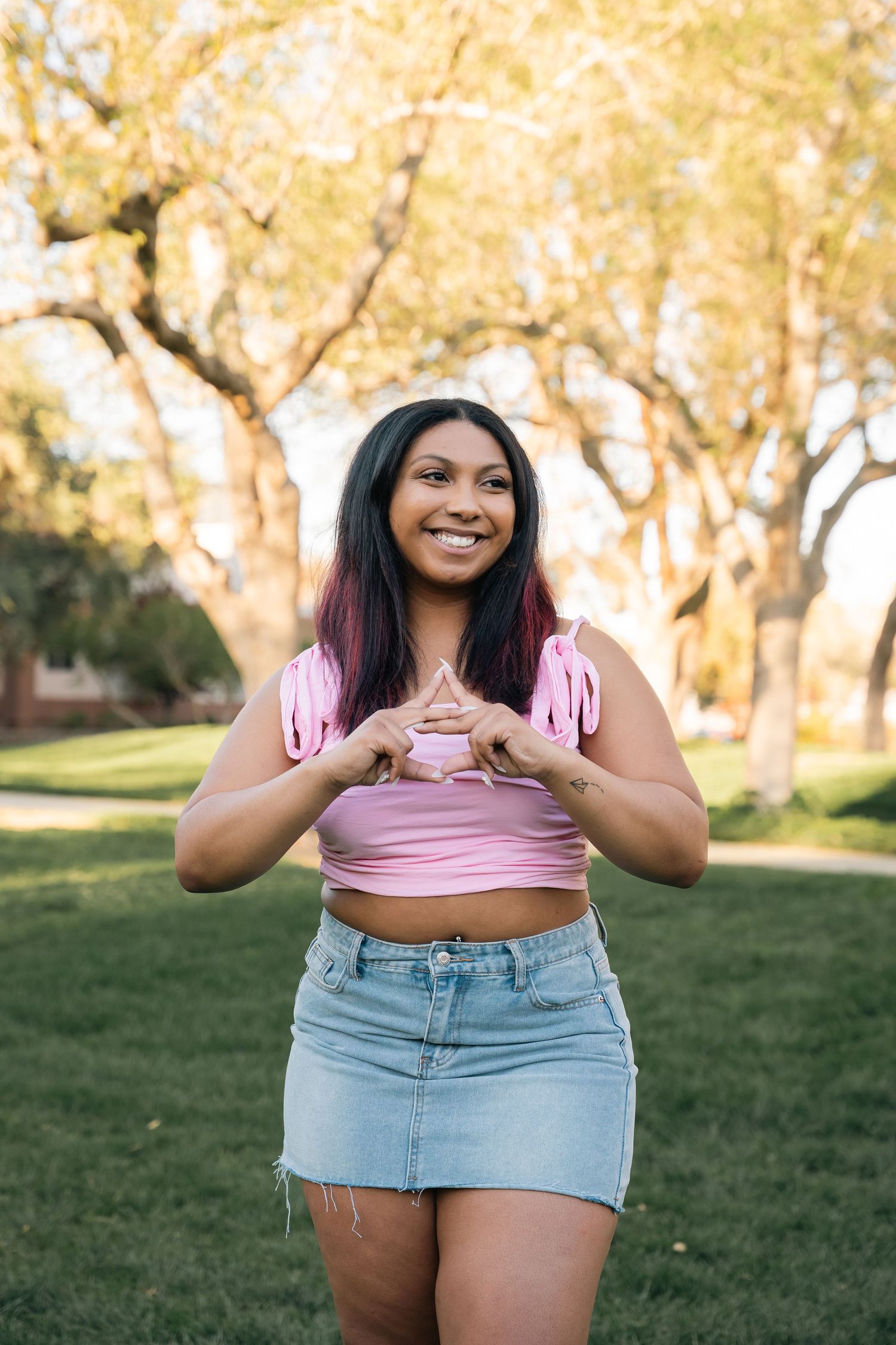 Woman in pink crop top and denim skirt poses outdoors with autumn trees in background.