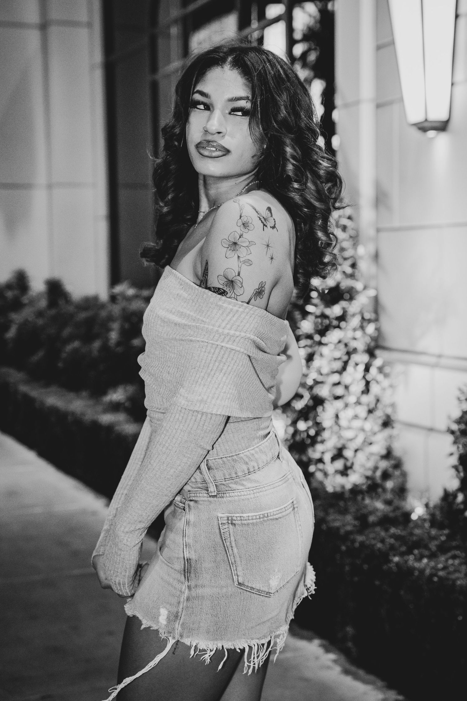 Monochrome portrait highlighting shoulder tattoo and styled wavy hair.