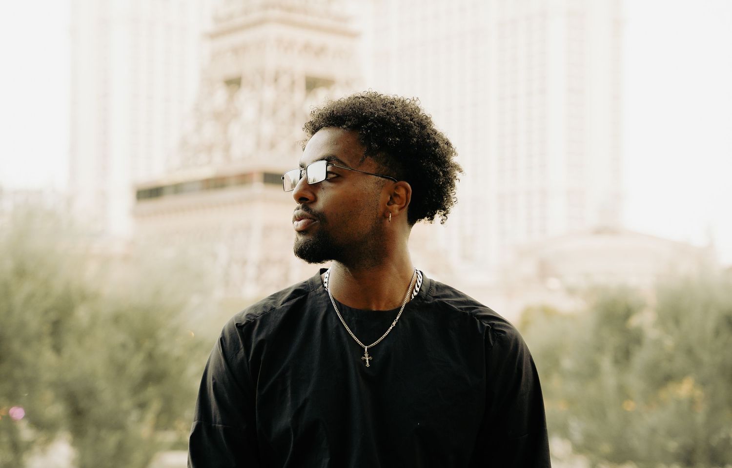 Stylish portrait of person in black wearing sunglasses against a blurred architectural backdrop.