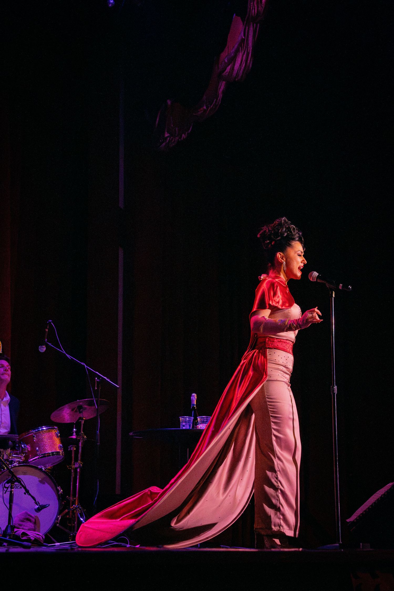A performer in a flowing red dress sings on stage with dramatic lighting and a drum kit visible.