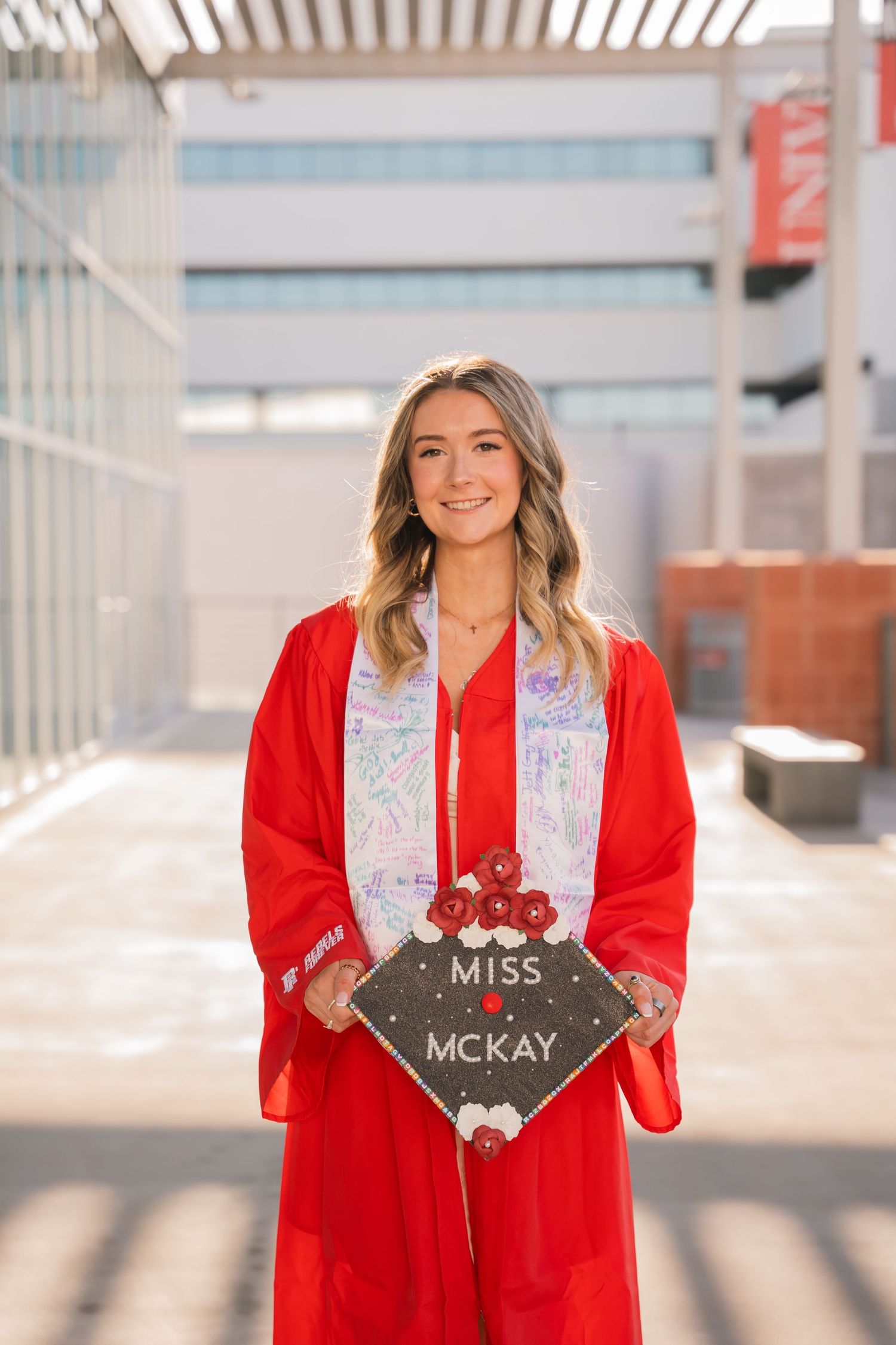 Graduate in red gown holds decorated mortarboard cap during campus photo session.