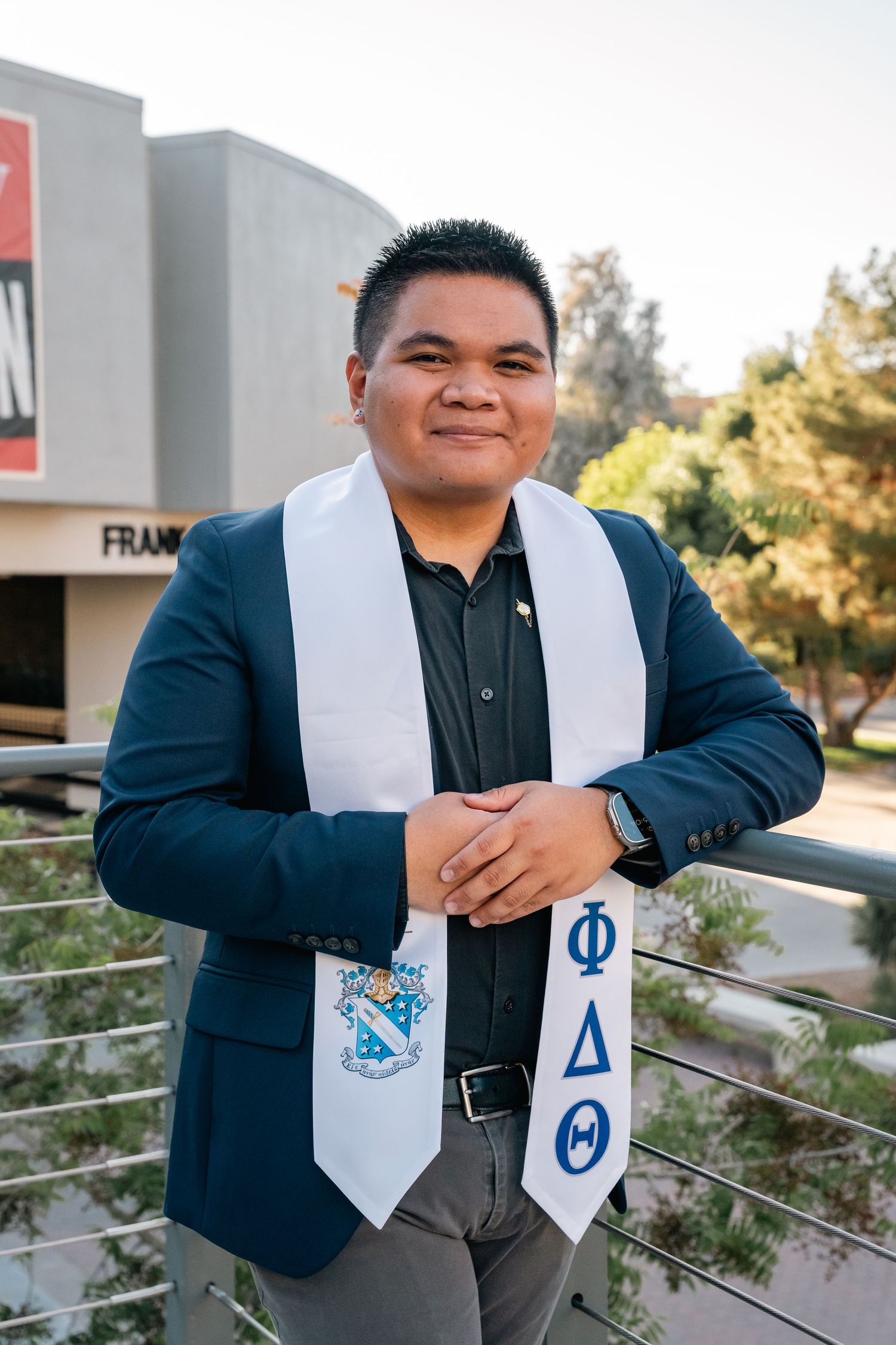 Graduate in navy blazer displays fraternity stoles while posing outdoors.