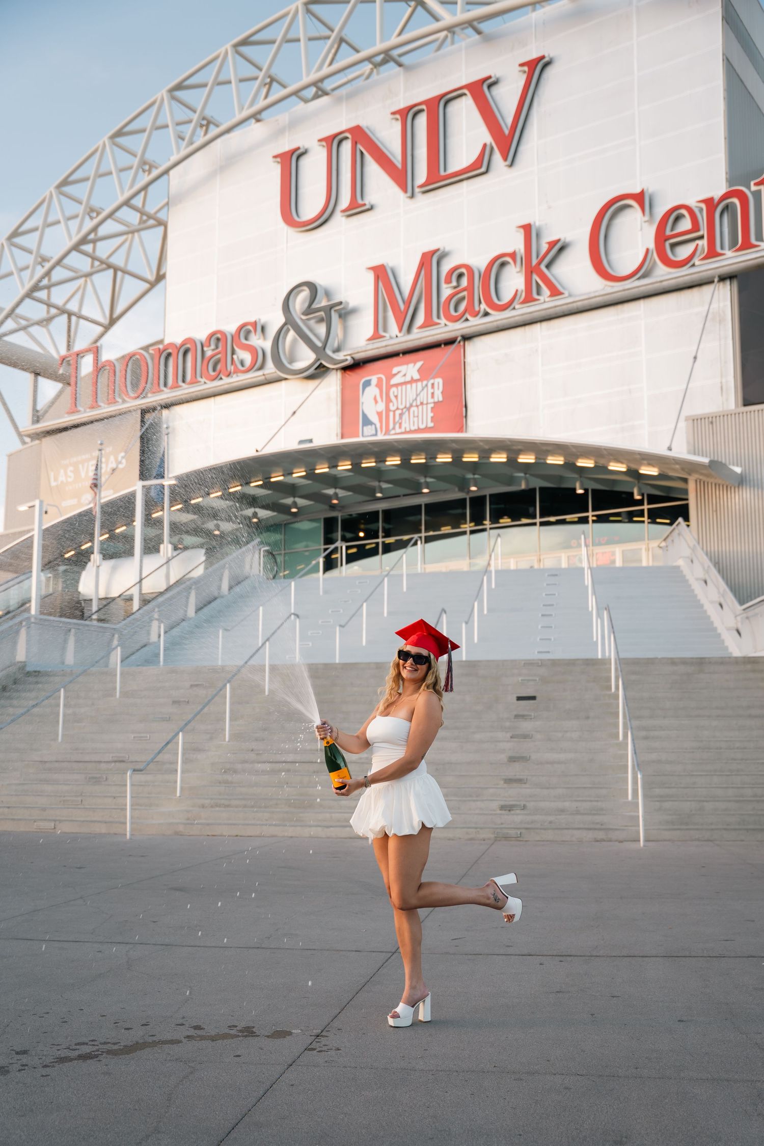 Graduate in white dress performing celebratory poses at UNLV campus steps.