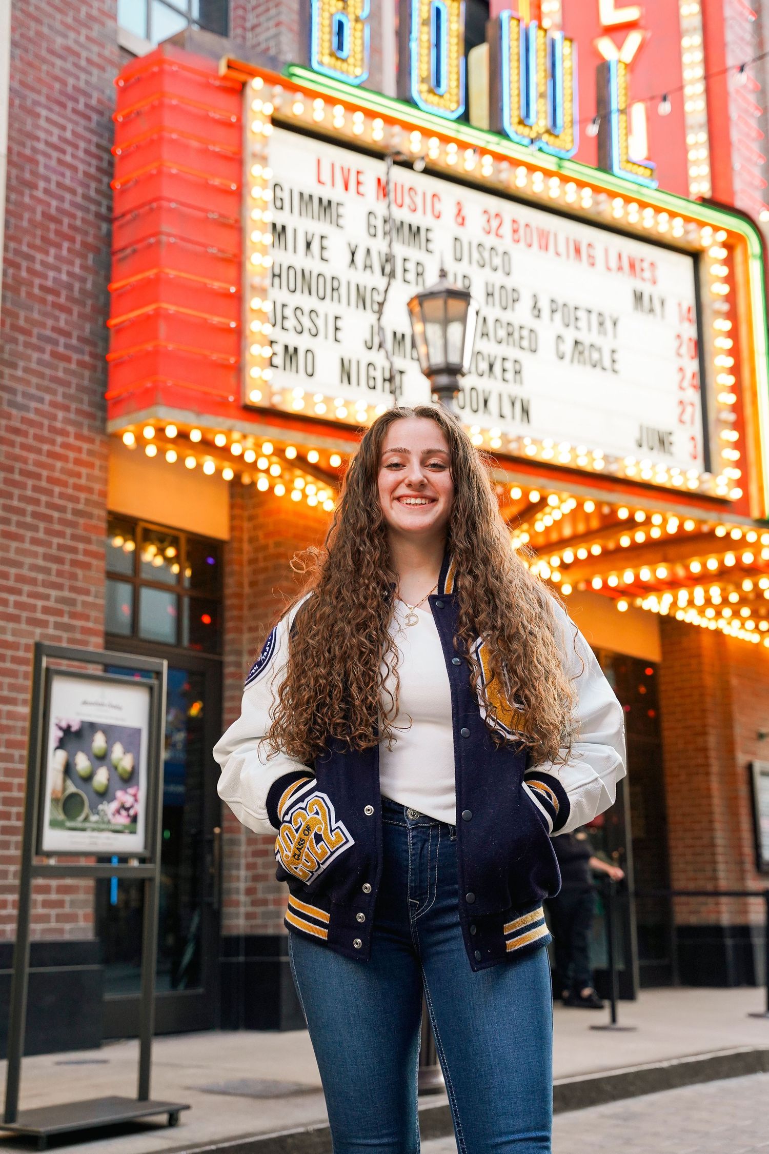 Student in letterman jacket poses in front of illuminated movie theater marquee.