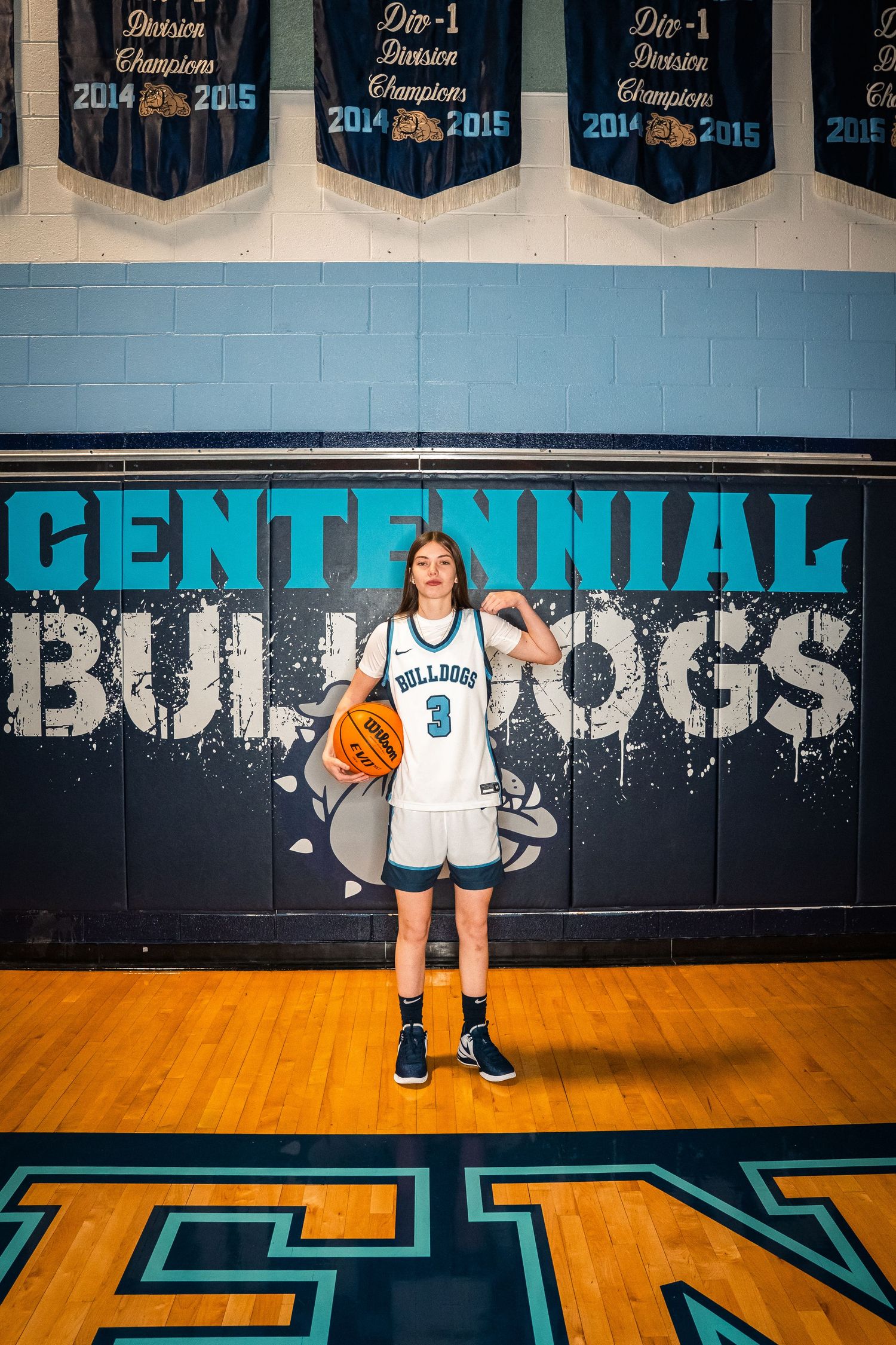 Basketball player in white jersey holding orange ball in front of blue Centennial Bulldogs wall.