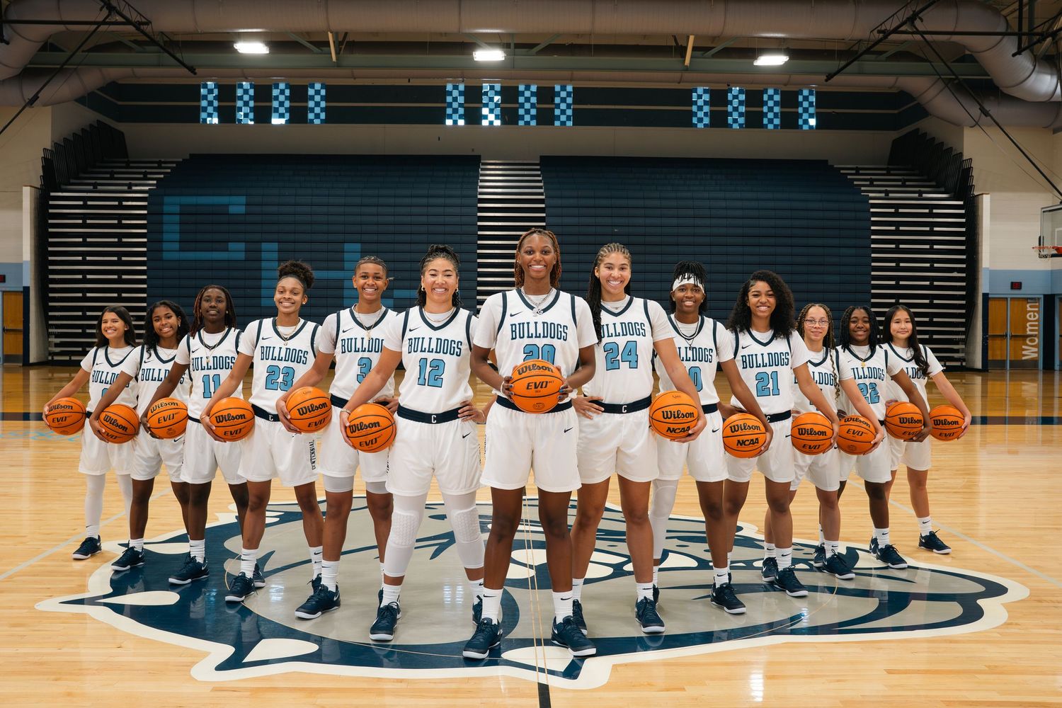 Basketball team lined up on court holding basketballs in white uniforms.