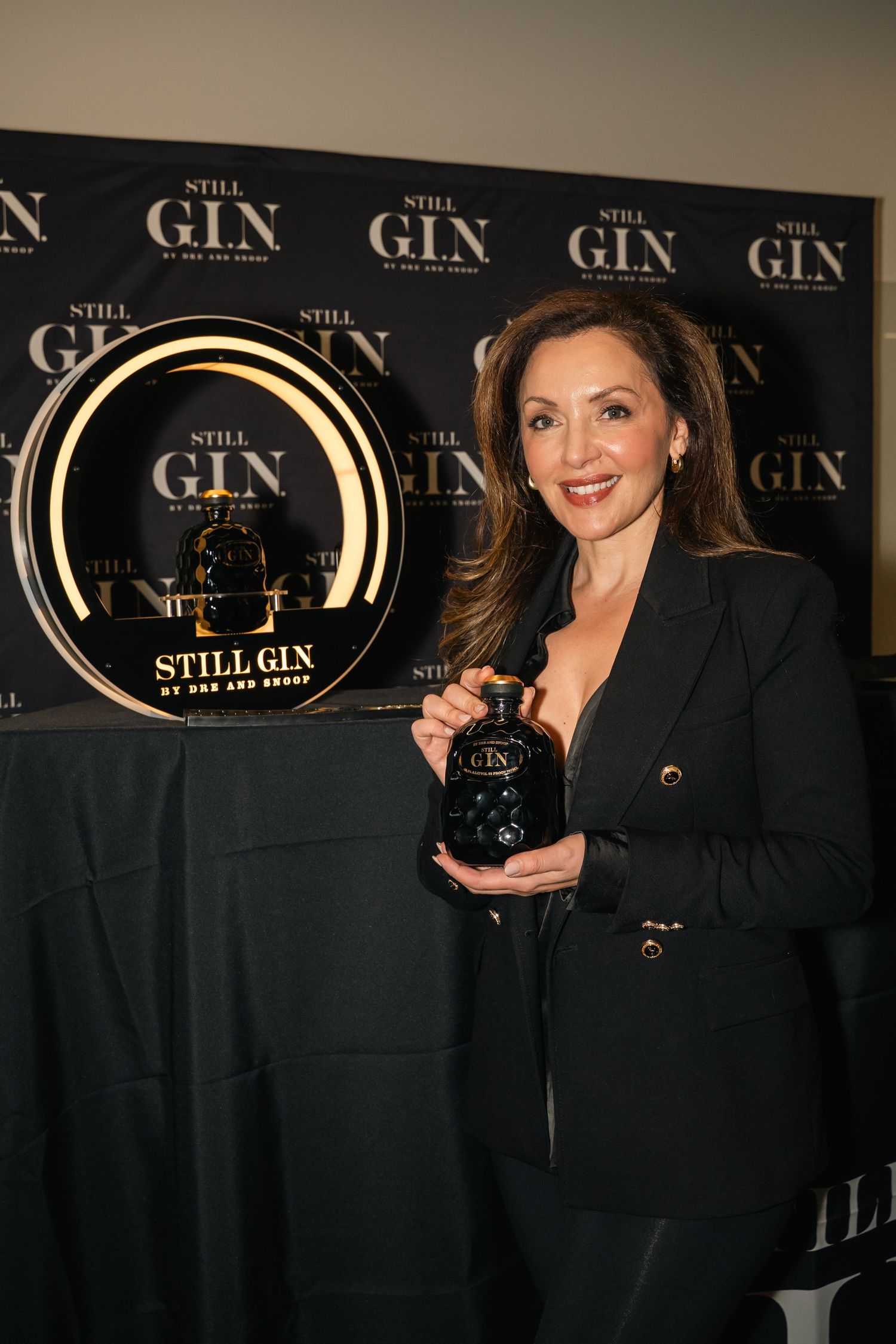 Person in black blazer posing with bottle against branded backdrop at gin promotion event.