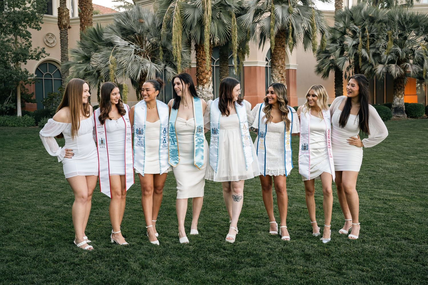 Group wearing coordinated white outfits and sashes stands together on lawn with palm trees.