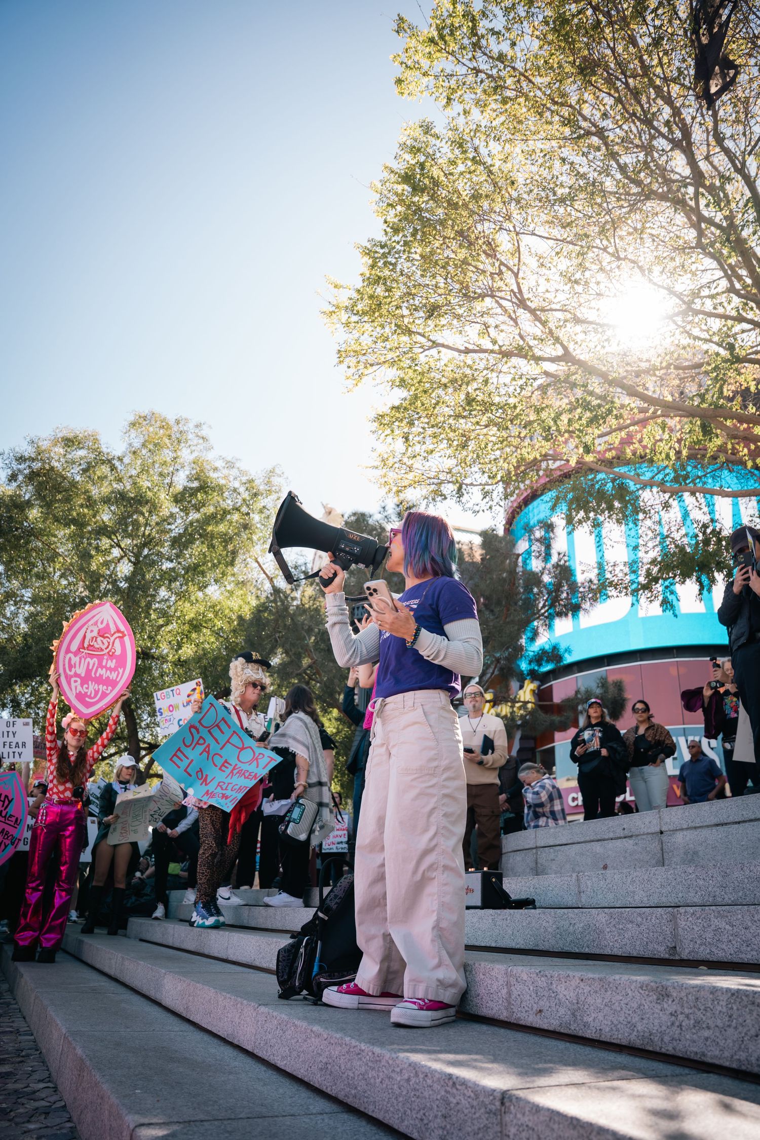 Person with a megaphone addresses crowd from steps during outdoor protest rally.