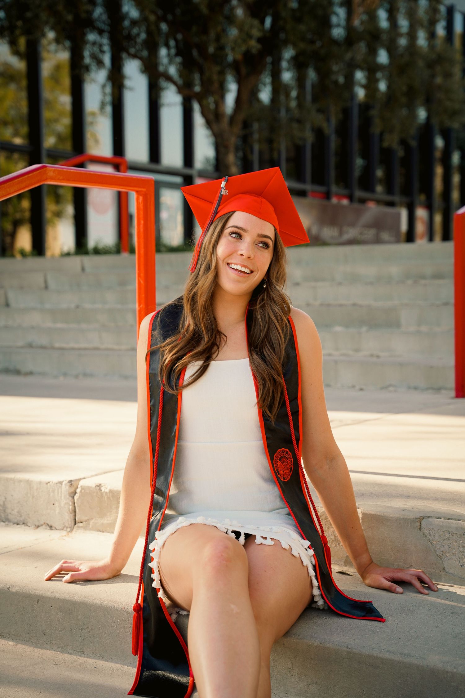 Monaco Media. Graduate in red cap and gown sitting on outdoor concrete steps during sunny graduation photoshoot.