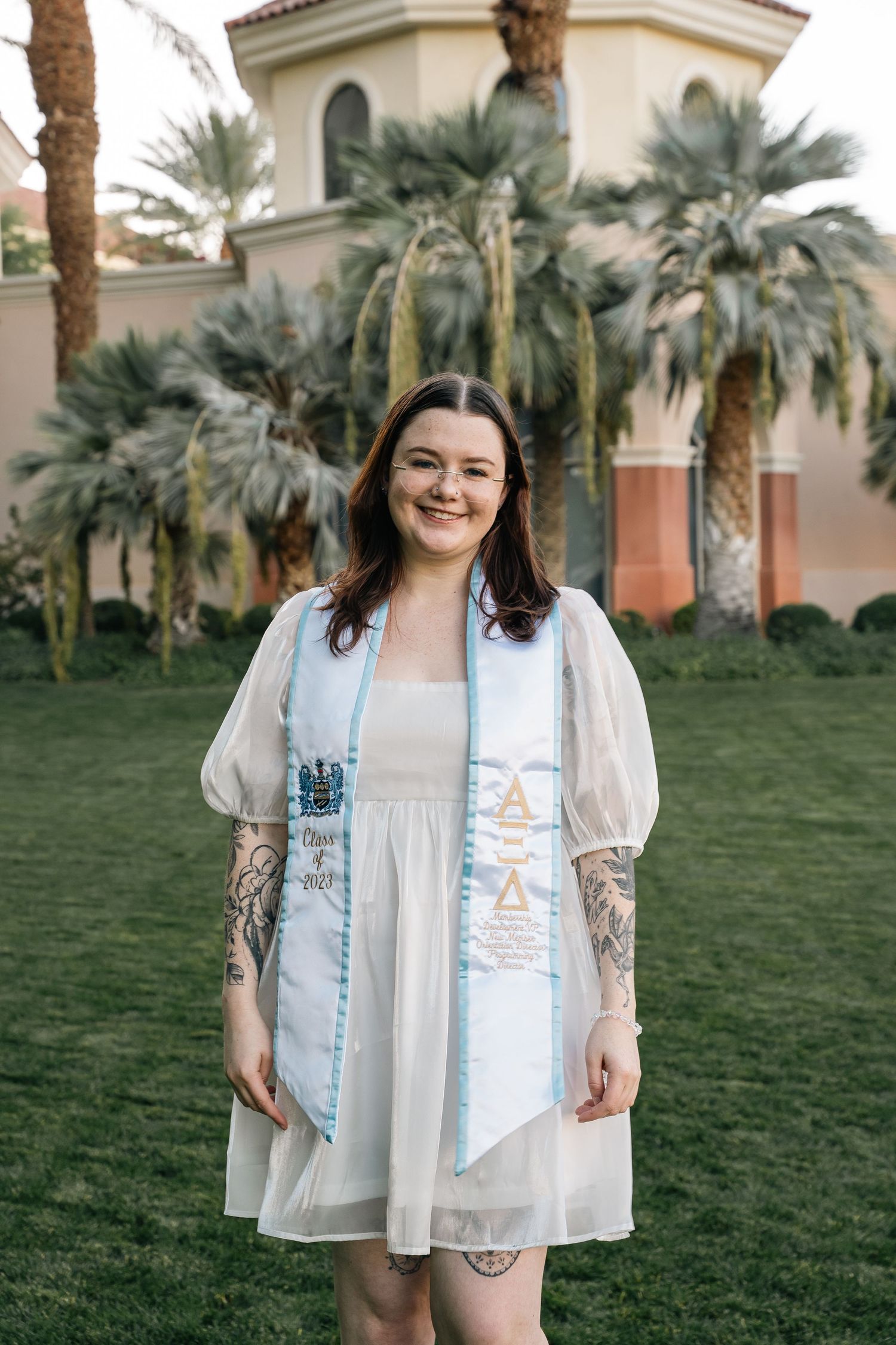 Graduate wearing white dress with puff sleeves and blue stole poses by palm trees and building.