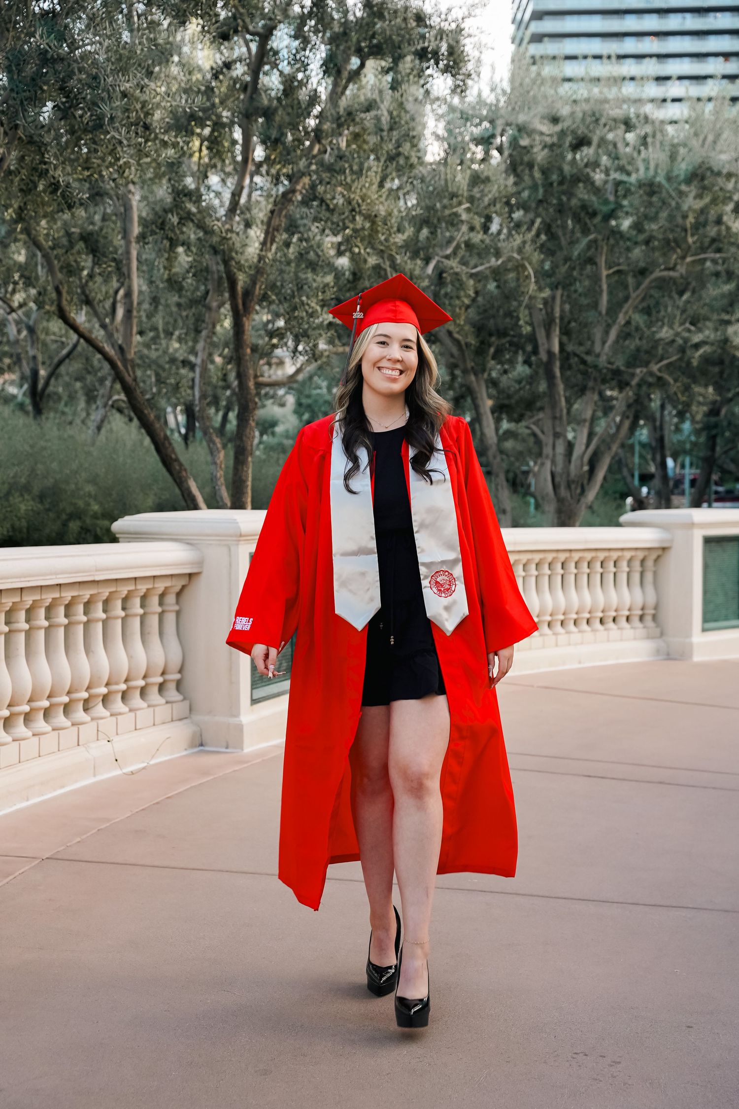 Graduate walks in red cap and gown along outdoor terrace with urban landscape behind.