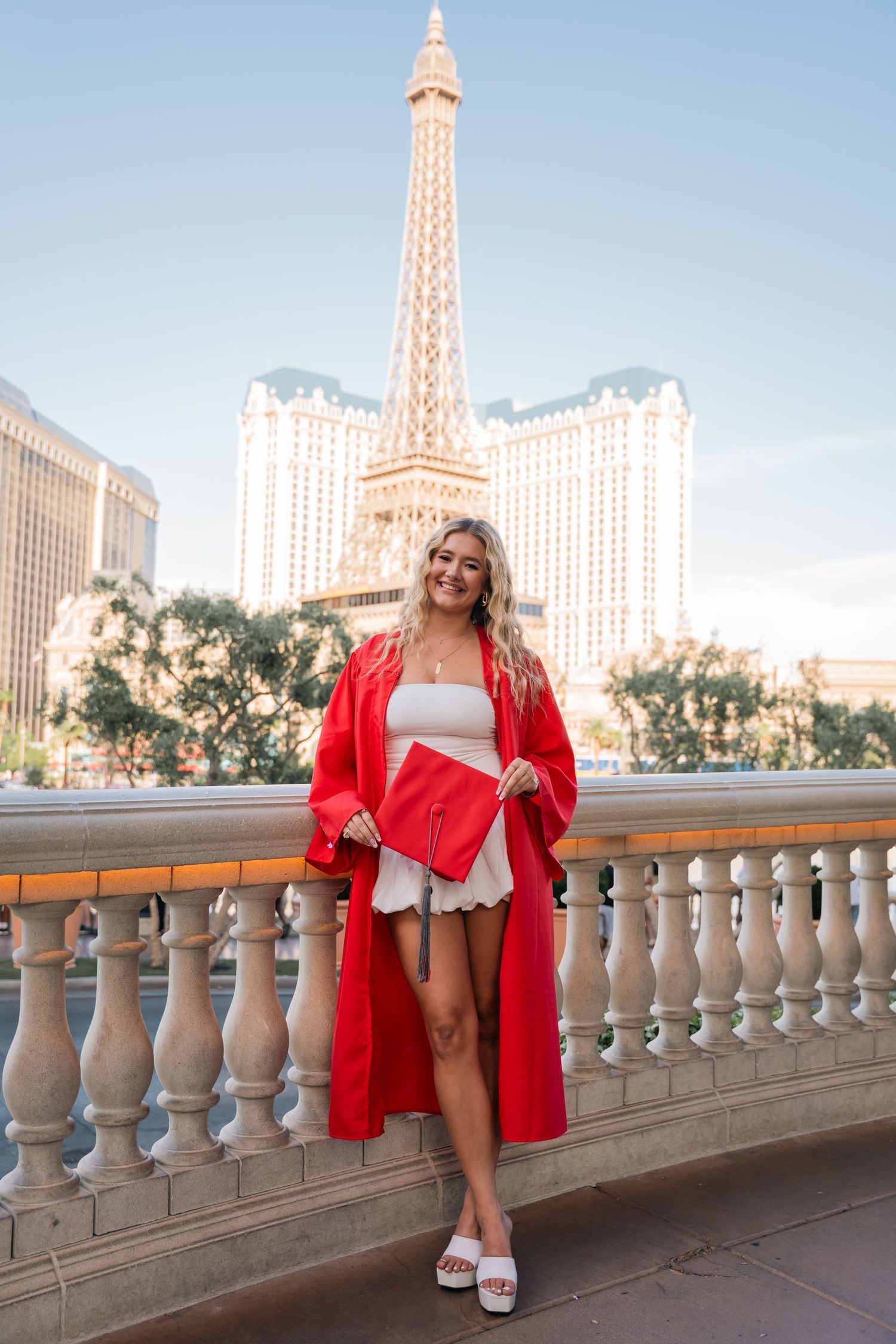 Graduate poses in red cap and gown by balcony railing with Las Vegas Strip and Eiffel Tower replica in background.