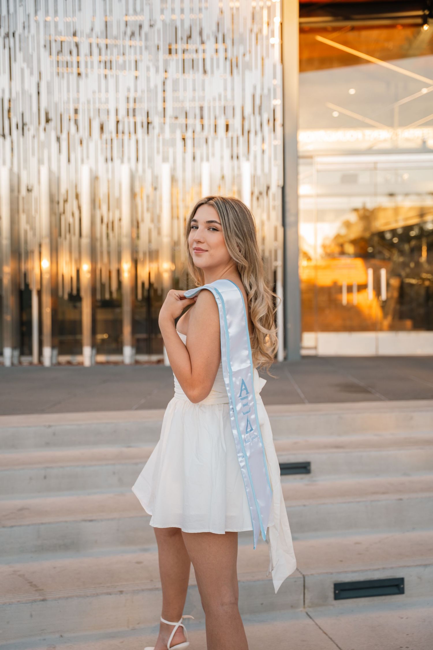 Graduate in white dress poses against metallic wall backdrop near modern building entrance.