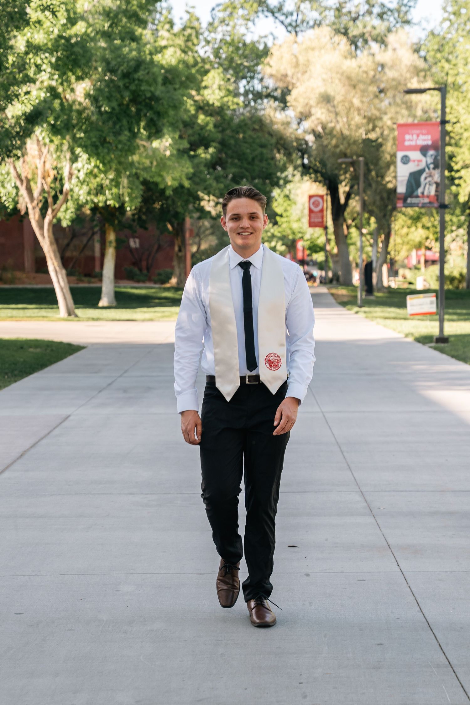 Graduate in professional attire walks confidently down campus pathway with trees in background.