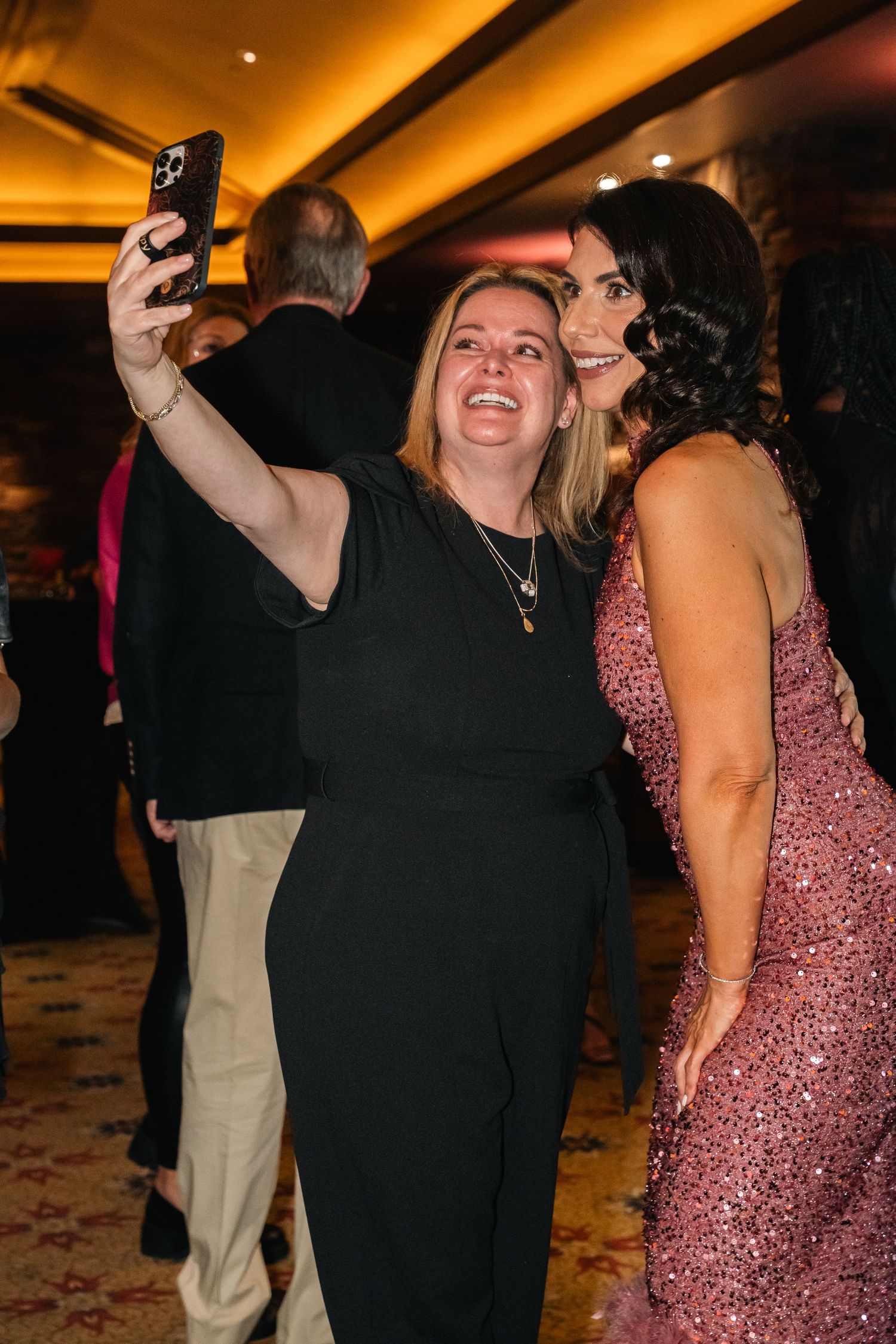 Two people taking selfie at formal event with warm lighting.