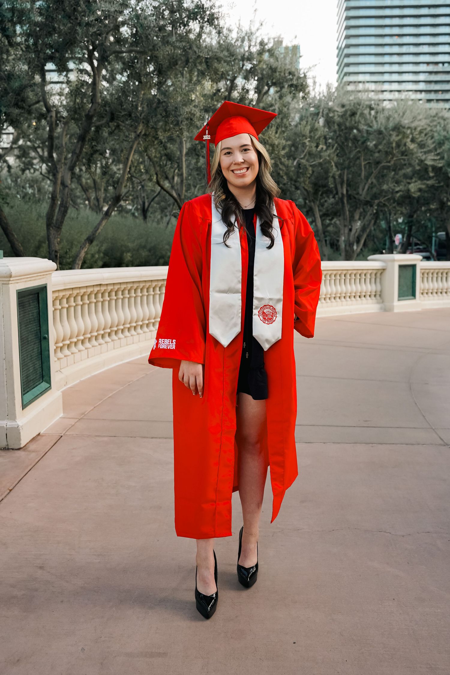 Graduate poses in bright red cap and gown with white stole on outdoor terrace with city buildings behind.