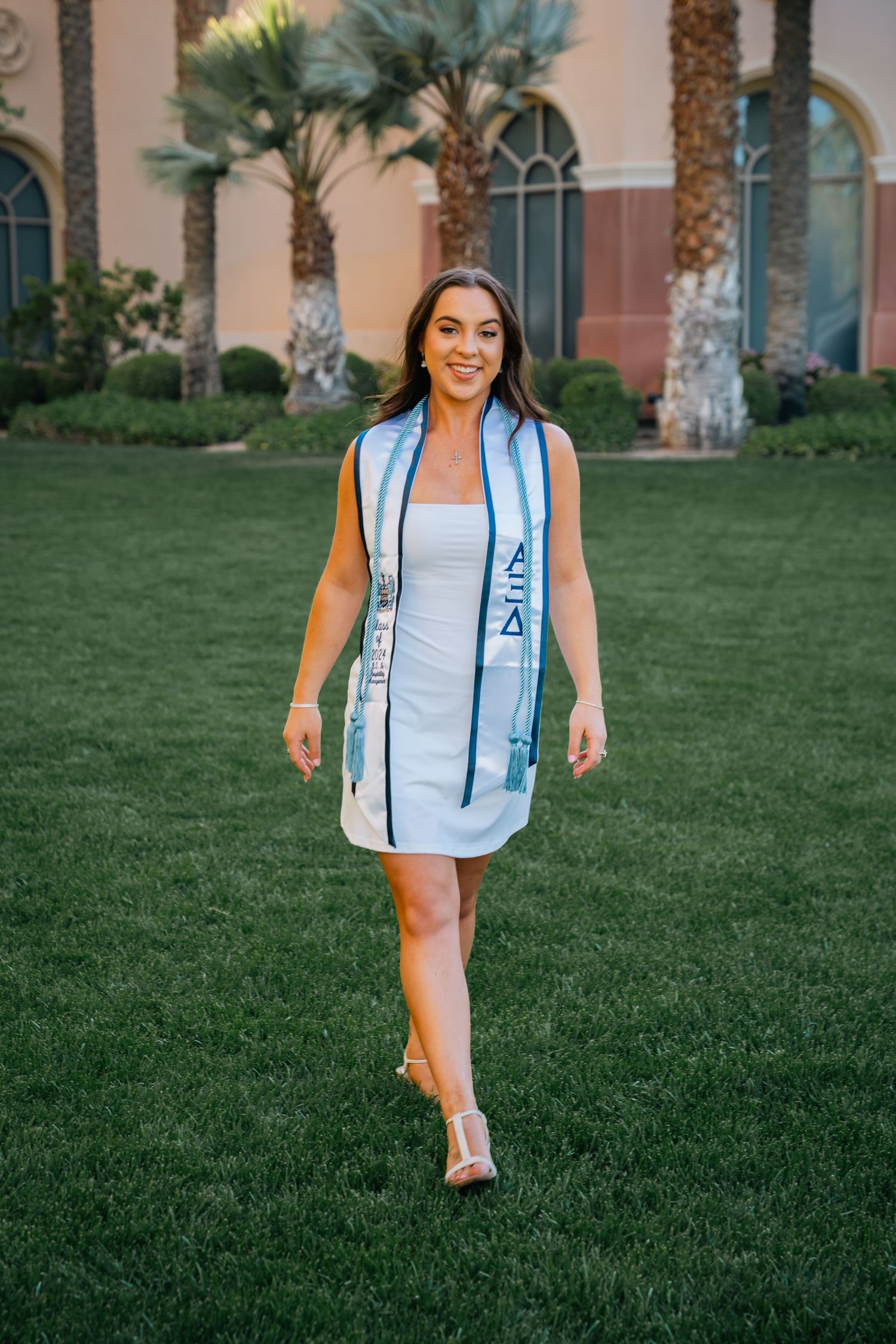 Graduate wearing white dress and blue stole walks across grass with palm trees in background.