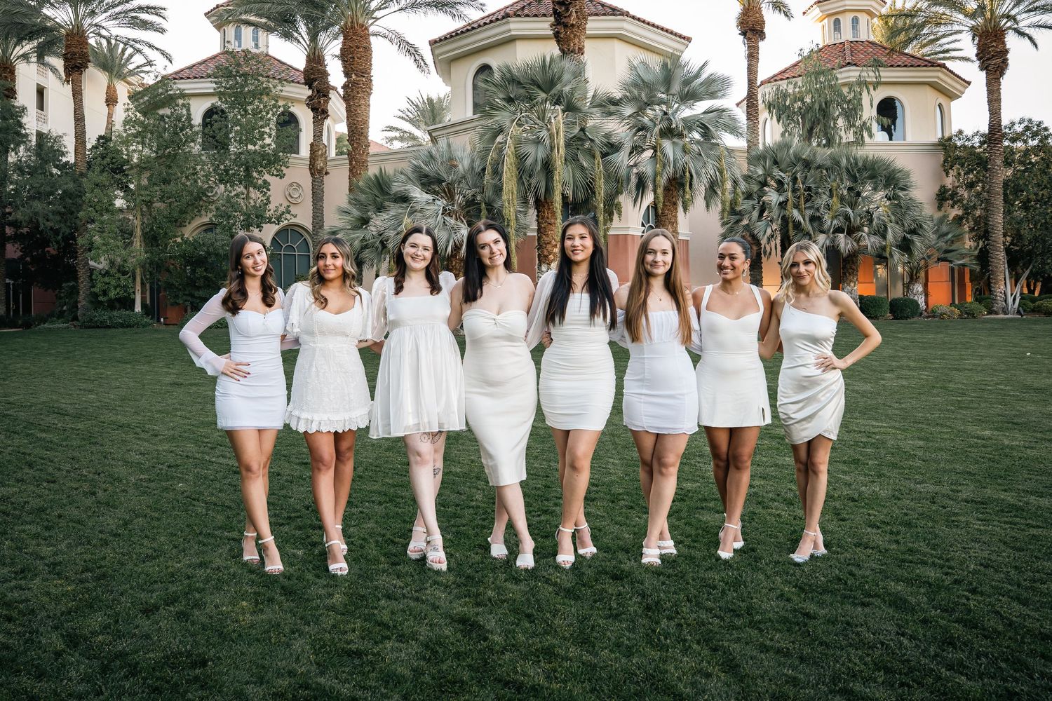 Row of graduates in white dresses pose on lawn in front of Spanish colonial style building.