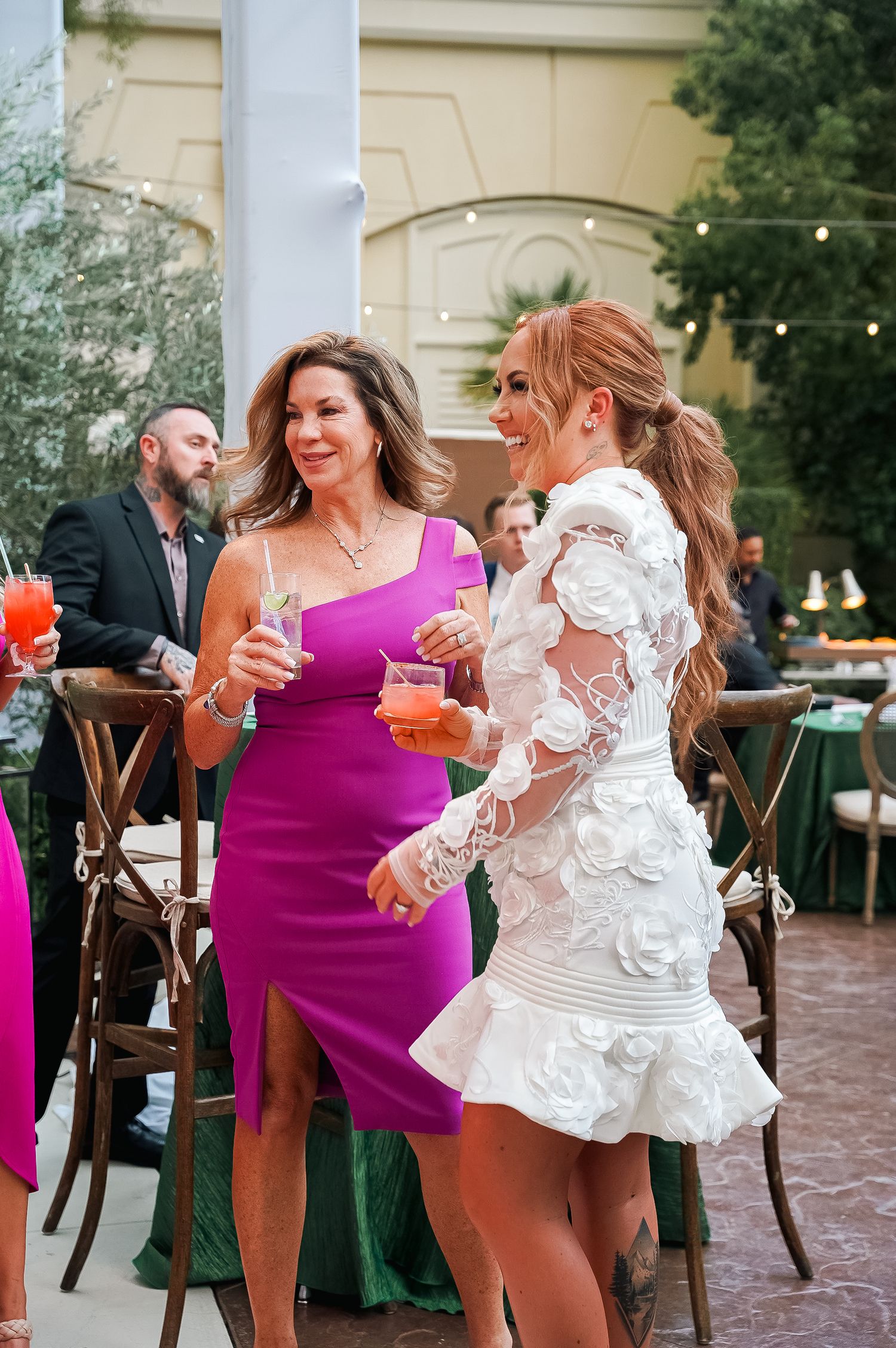 Two guests in cocktail attire share a laugh while enjoying drinks at an outdoor party.
