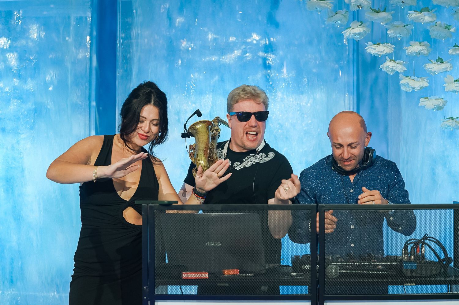 Three musicians performing together at an event with blue backdrop and technical equipment.