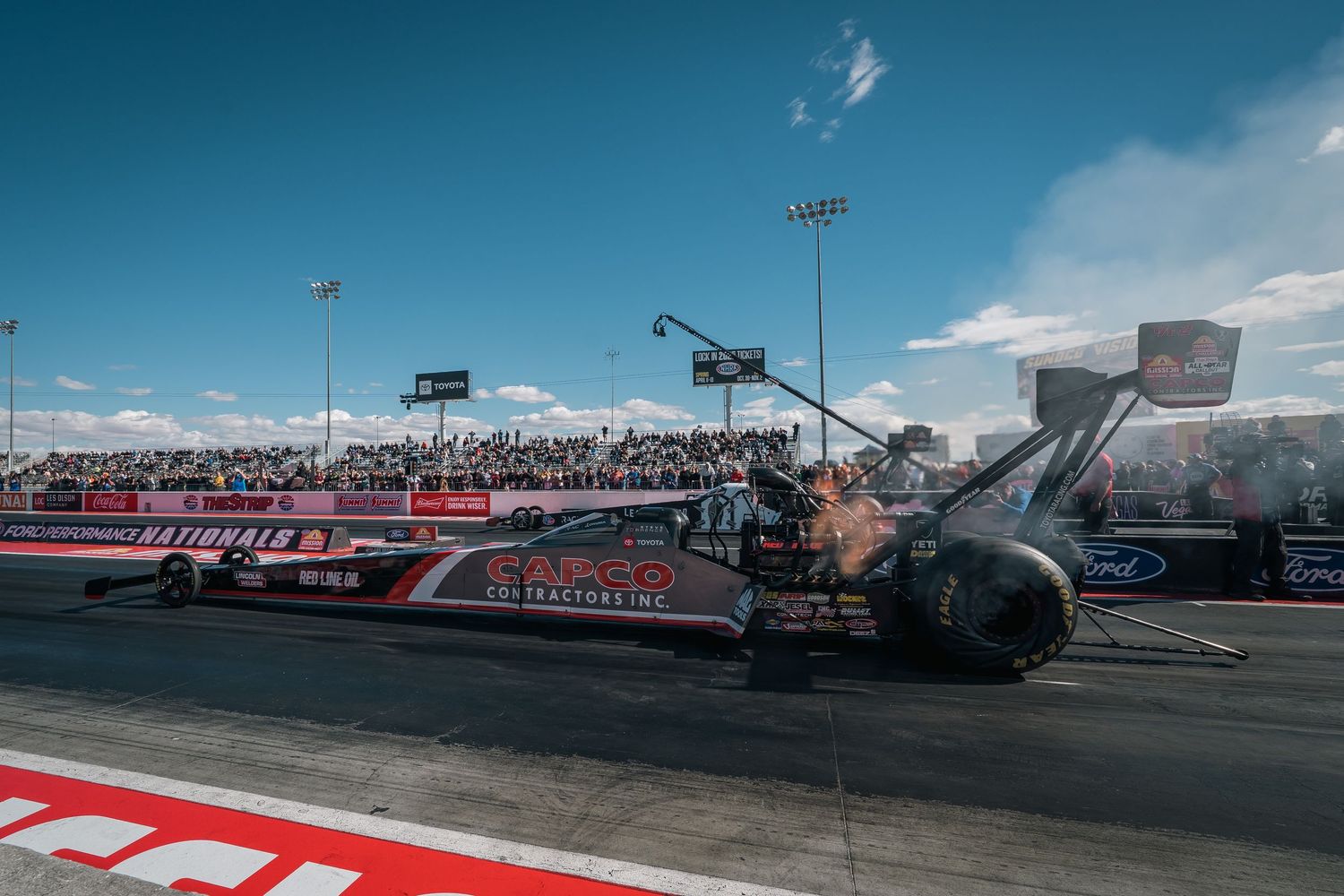 Top fuel dragster racing down a drag strip with spectator stands visible in the background.