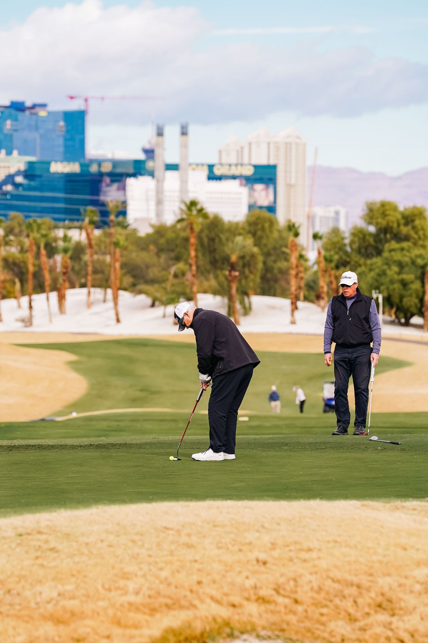 Two golfers practicing putting on a desert golf course with city buildings in the background.