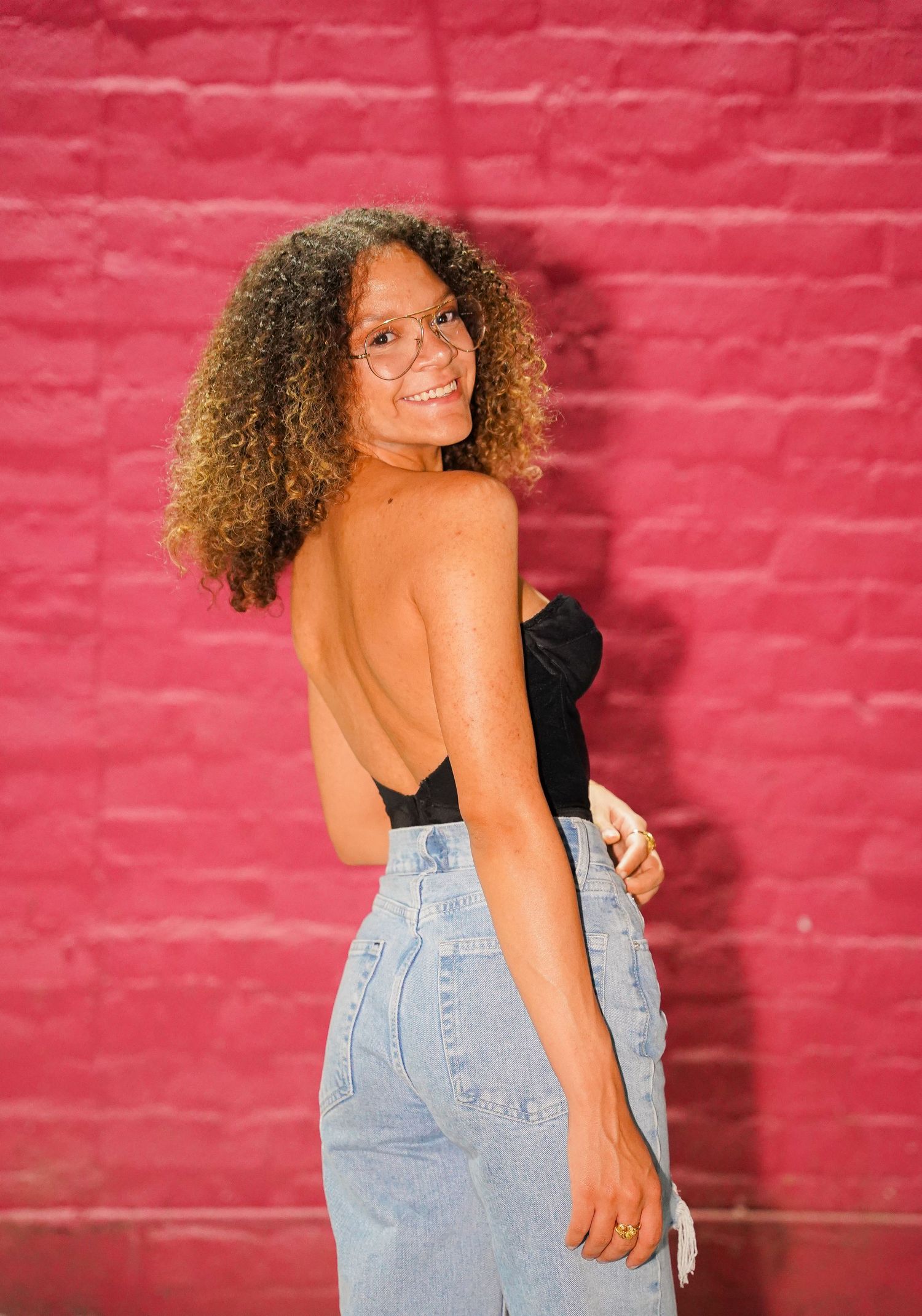 A person wearing a black top and light blue jeans poses against a pink brick wall background.