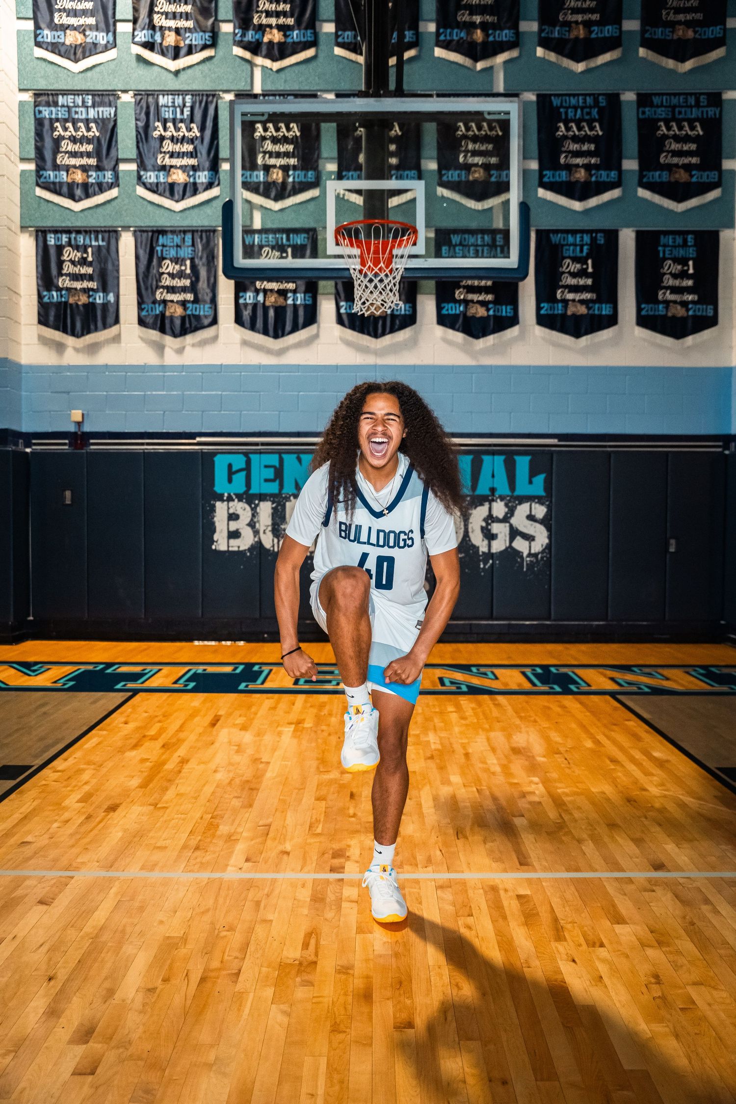Basketball player jumps energetically on court with Centennial Bulldogs banners and logo in background.