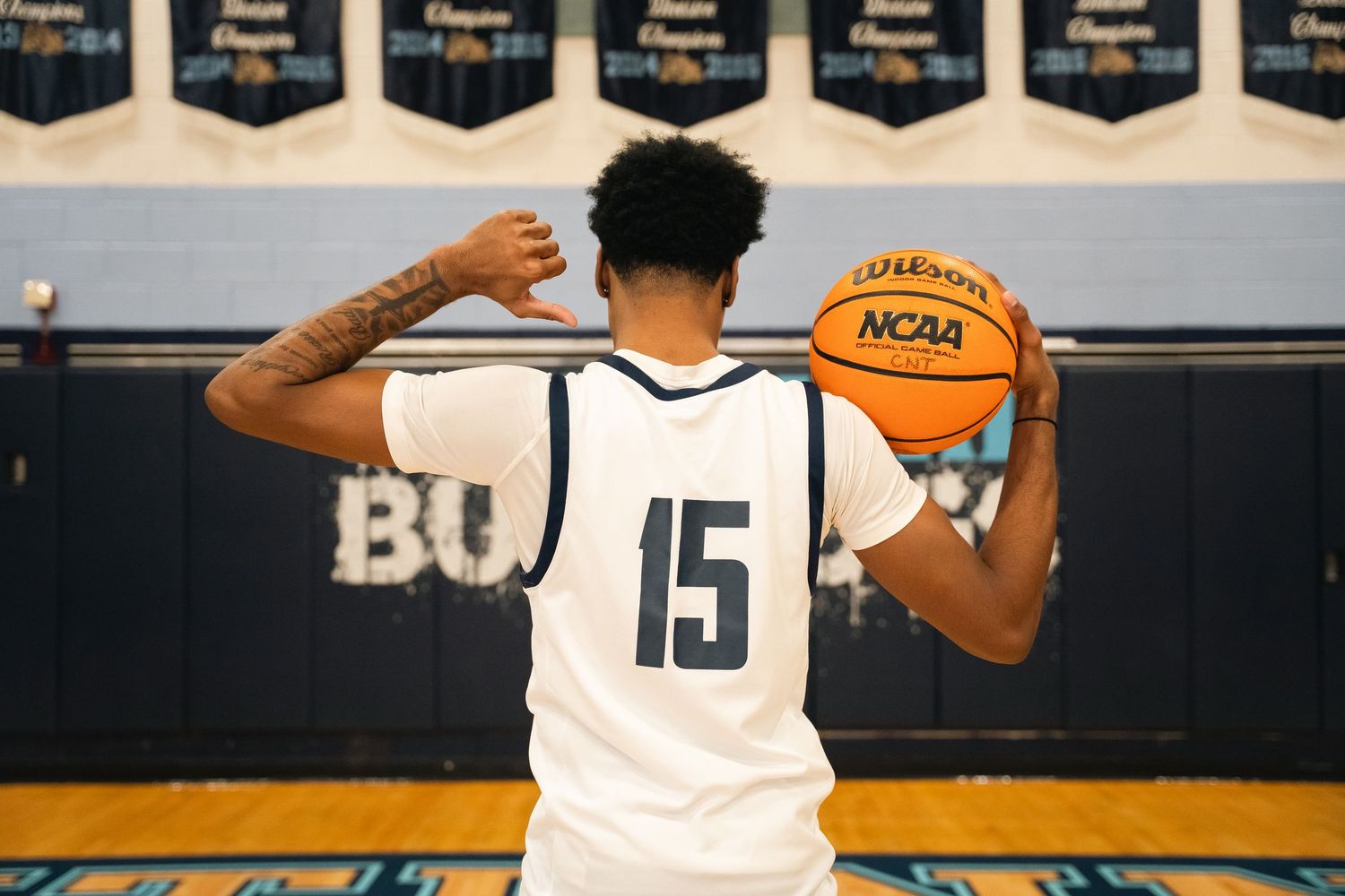 Basketball player wearing jersey number 15 holds ball while posing near championship banners.