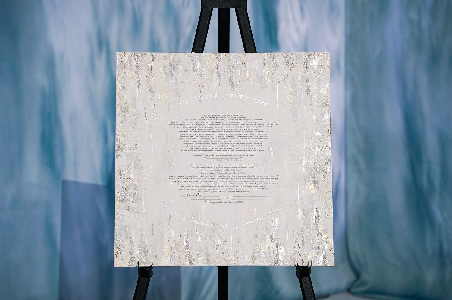 White decorative sign displayed on easel against blue-tinted backdrop at wedding venue.